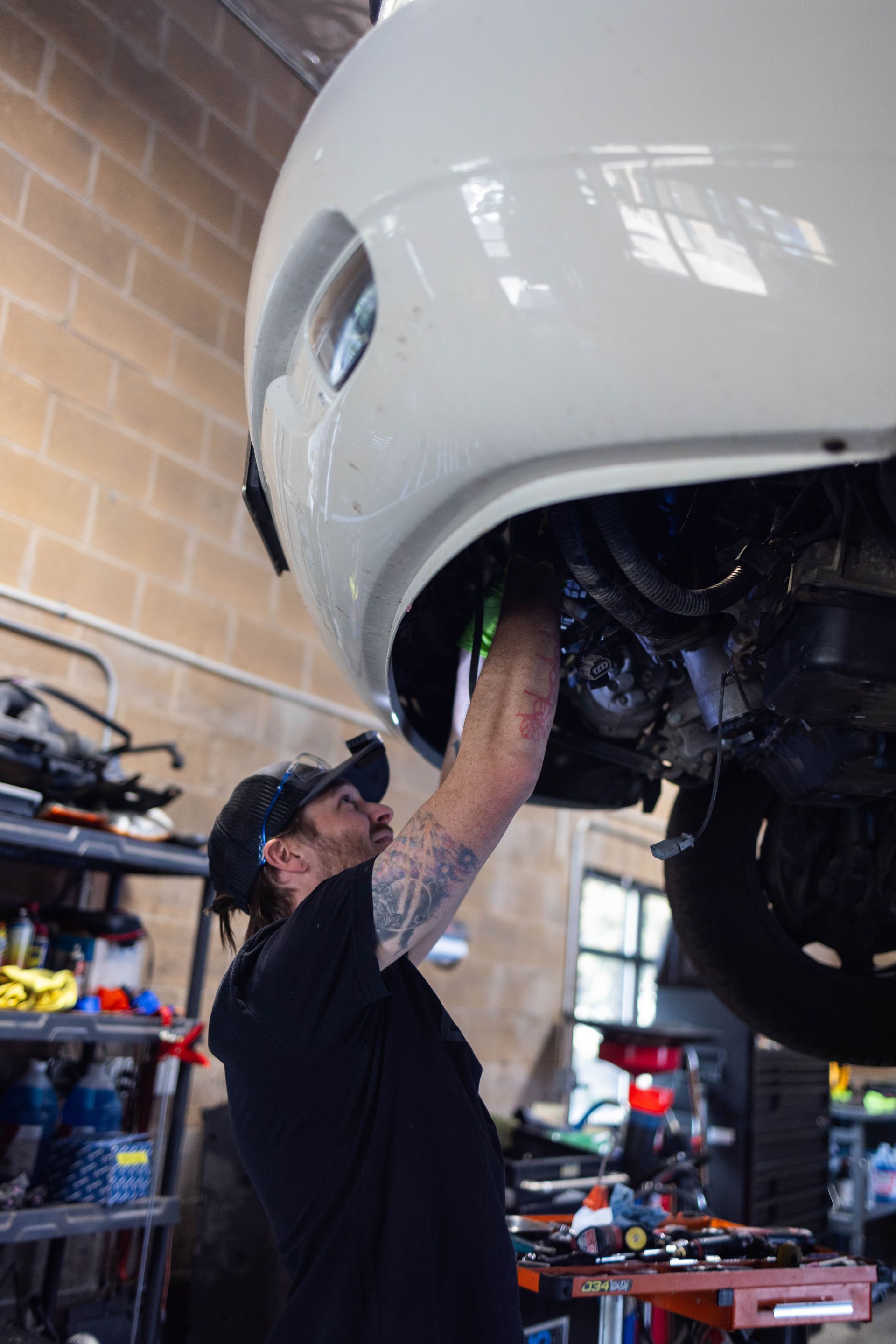 Mechanic working under a white car. He wears a black shirt and hat; a garage setting with tools. | Peak Euro