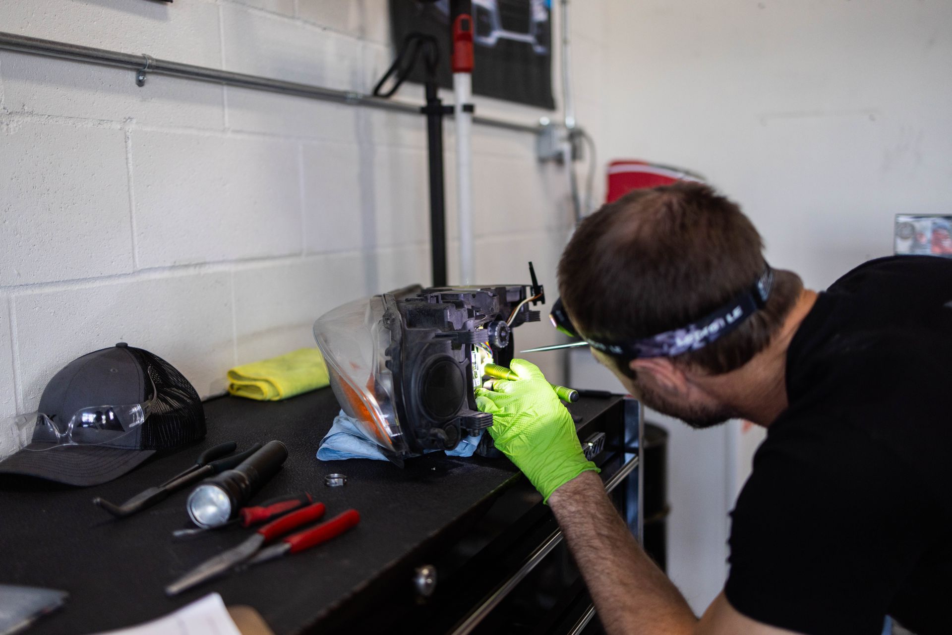 Person in a garage, working on a car headlight with tools, wearing glasses and gloves. | Peak Euro