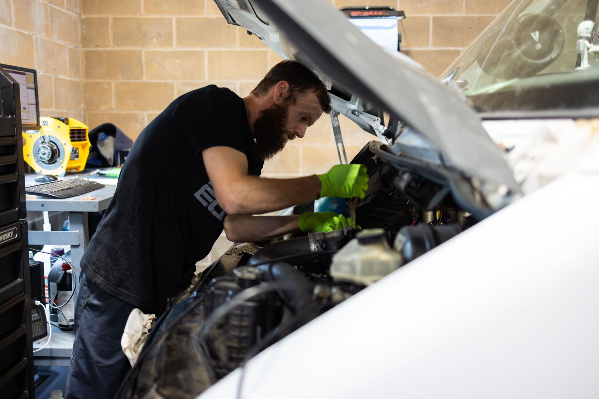 Mechanic with beard, green gloves, works on car engine in garage. | Peak Euro