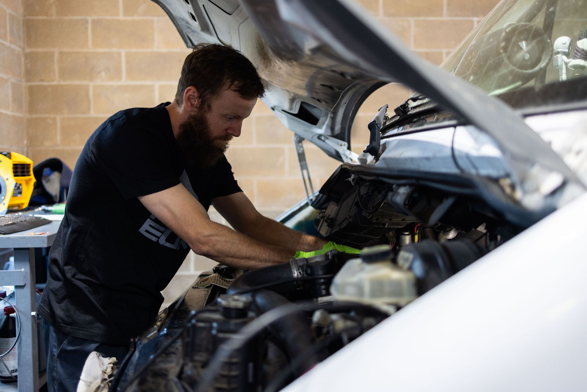 Mechanic with beard working on a car engine under the open hood in a garage. | Peak Euro