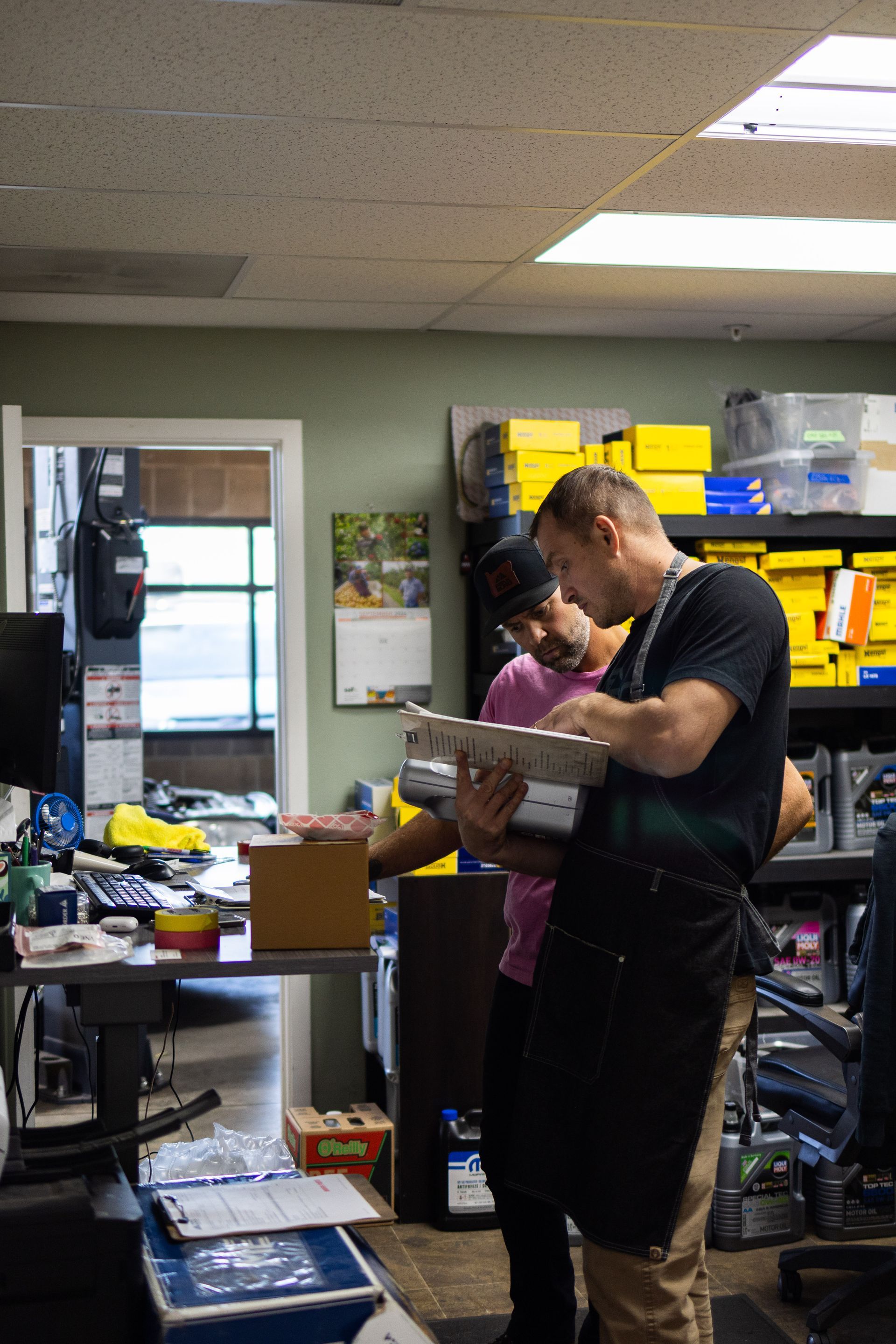 Two men in a cluttered workshop review paperwork. One wears a black apron, the other a pink shirt. | Peak Euro