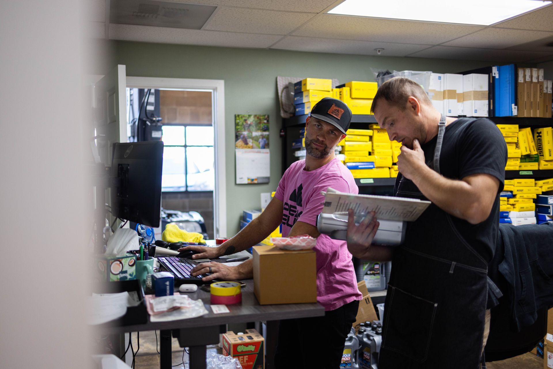 Two men reviewing paperwork in a cluttered office with boxes and supplies. One wears pink and the other black. | Peak Euro
