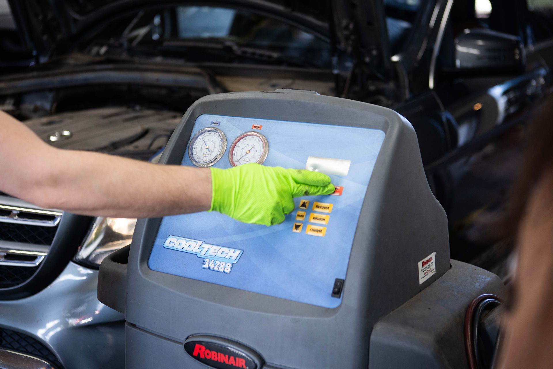 A technician with a green glove operates an air conditioning service machine in a car engine bay. | Peak Euro