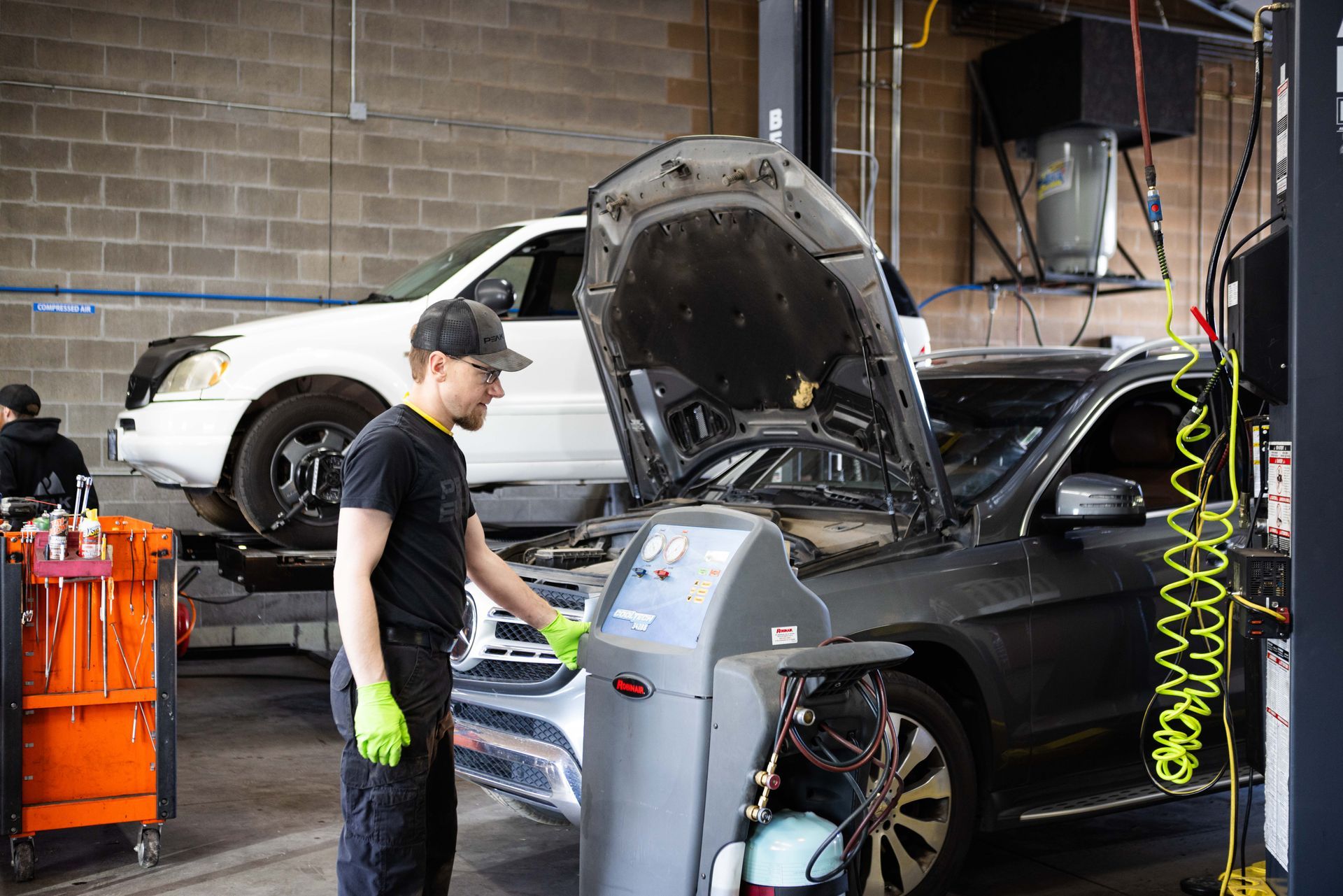 Mechanic working on a car with its hood open in an auto repair shop. Another car on a lift.  | Peak Euro