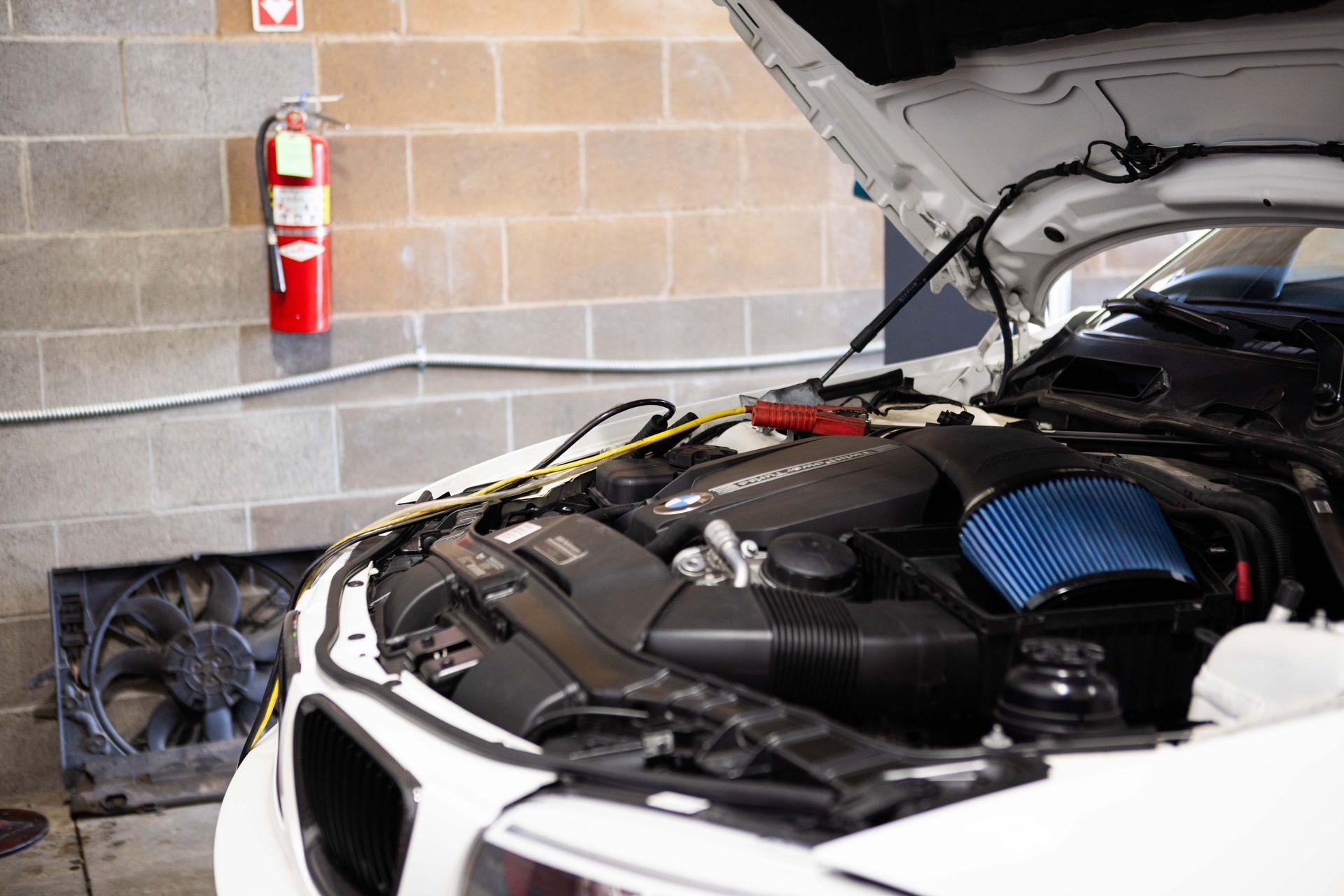 A white car's engine bay is open for repair in a garage, with a blue air filter and fire extinguisher. | Peak Euro