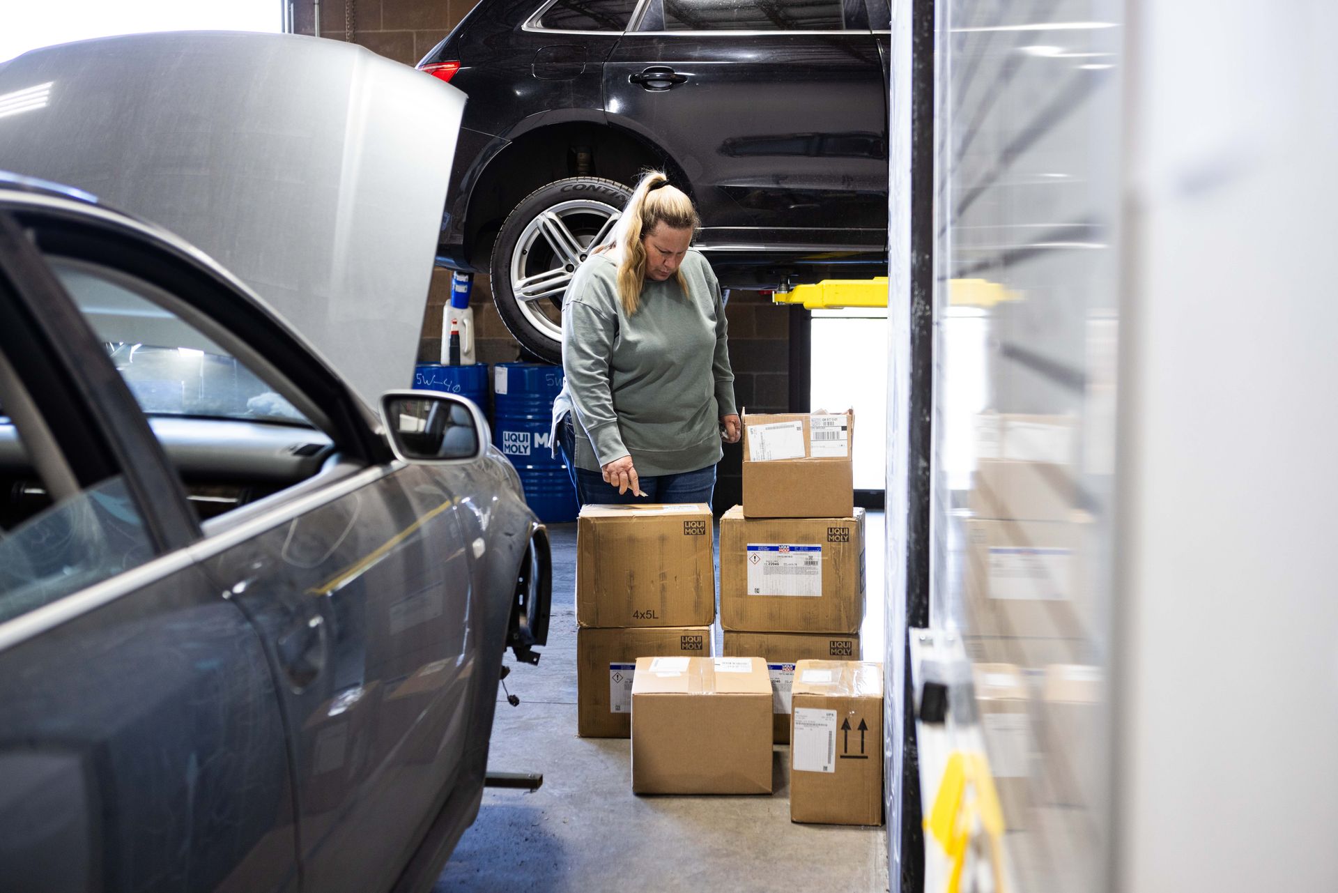Woman in auto repair shop examining boxes near shelving, another car on lift in background. | Peak Euro
