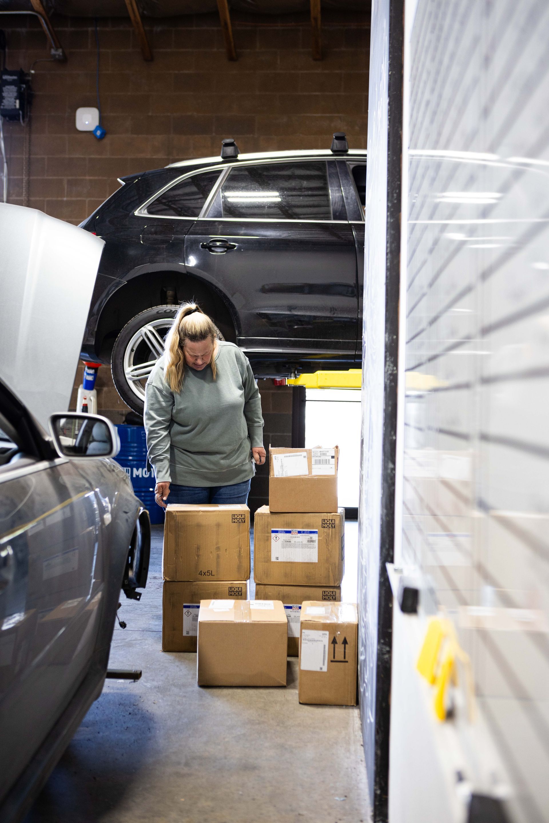 Woman in garage, looking at boxes stacked near a wall, with car on a lift in the background. | Peak Euro