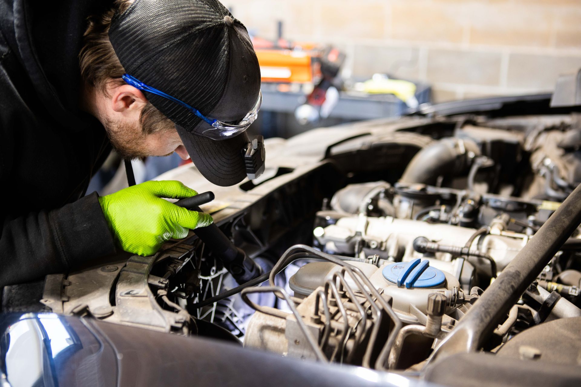 Mechanic examines car engine with a flashlight, wearing gloves, cap, and safety glasses in a garage. | Peak Euro