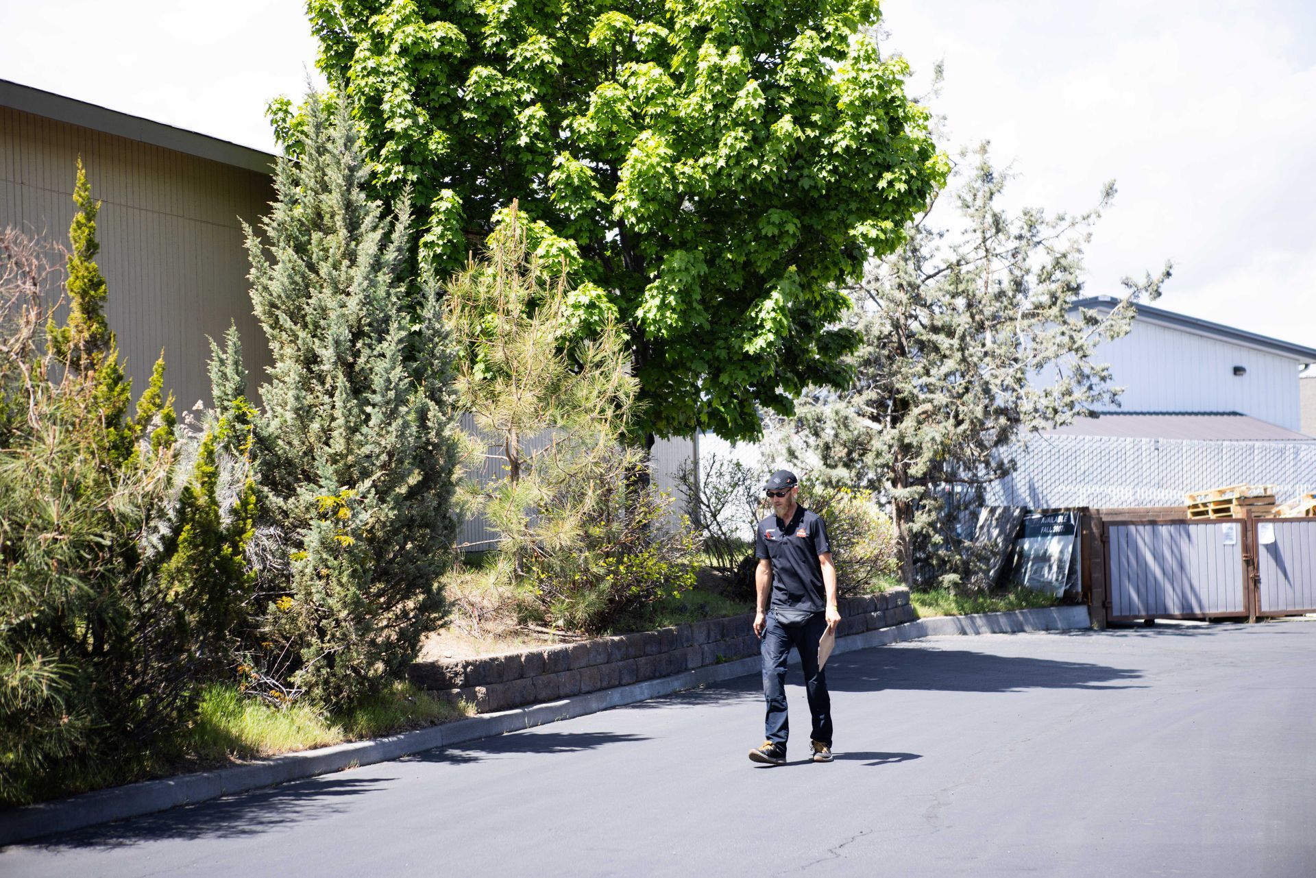 Man walking on a street, wearing a black shirt and jeans. Green trees and a white fence in background. | Peak Euro