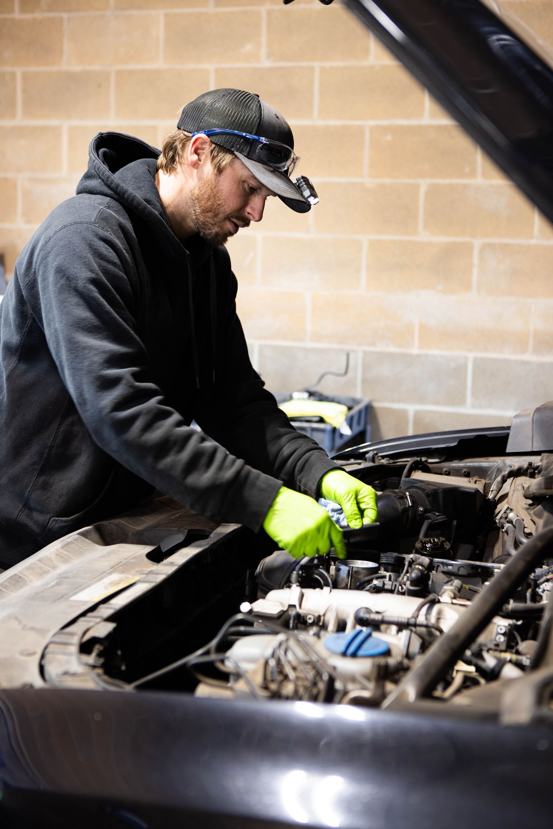 Mechanic working on a car engine in a garage, wearing gloves, hat, and headlamp. | Peak Euro