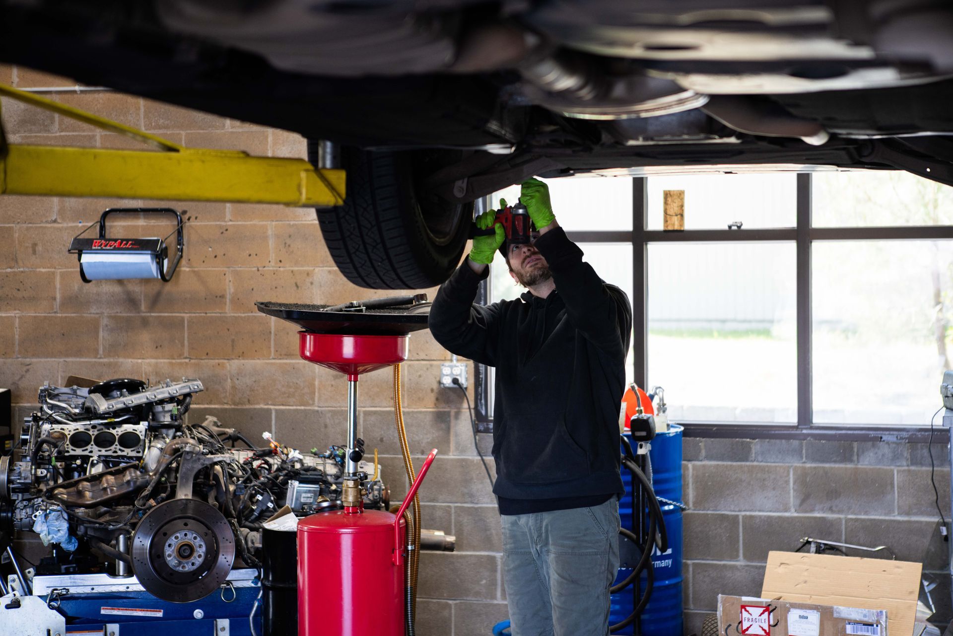 Mechanic working under a car lifted on a hoist in a garage, wearing gloves and a hooded sweatshirt. | Peak Euro