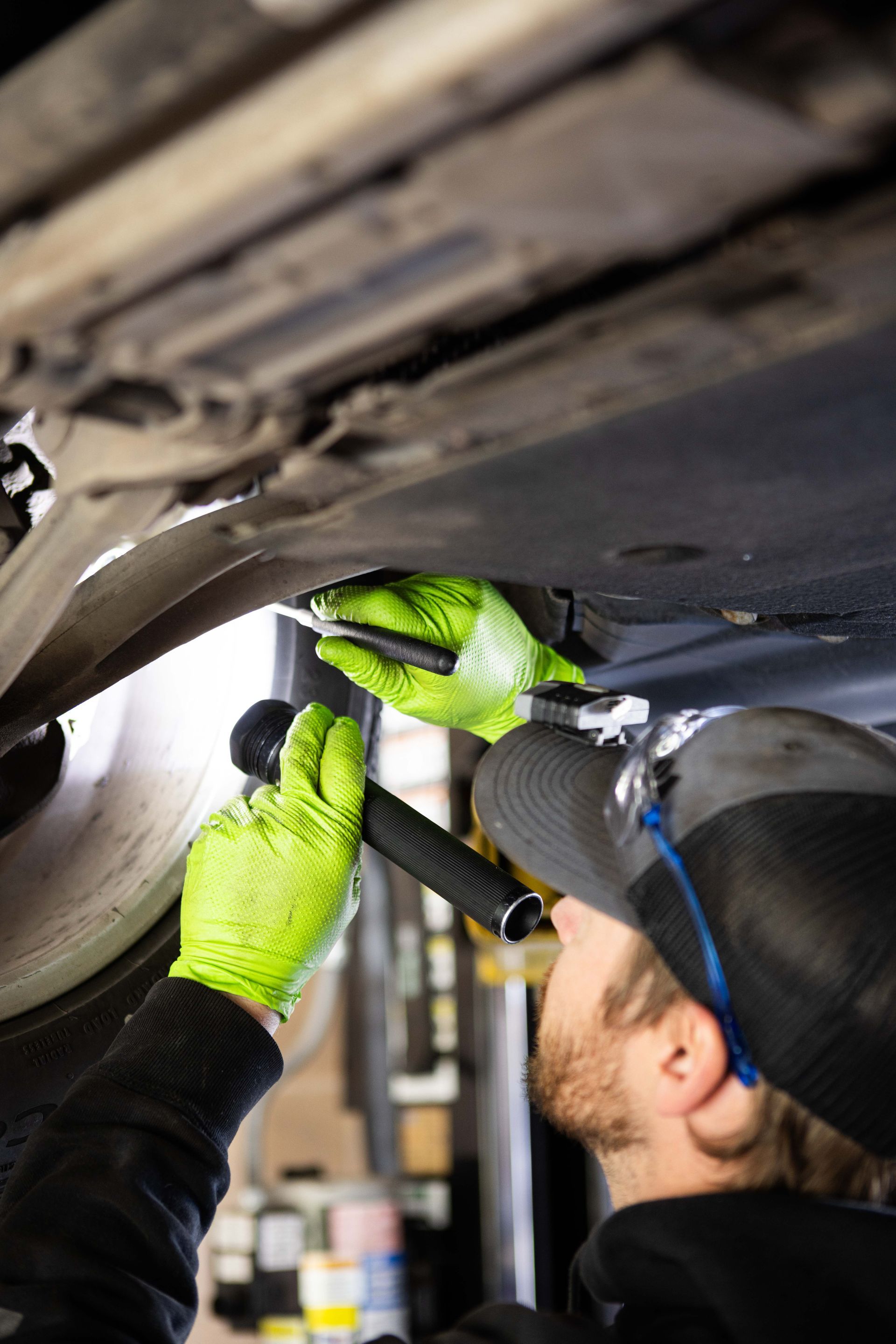 Mechanic under a car, wearing gloves, uses a flashlight and tool to inspect the vehicle. | Peak Euro