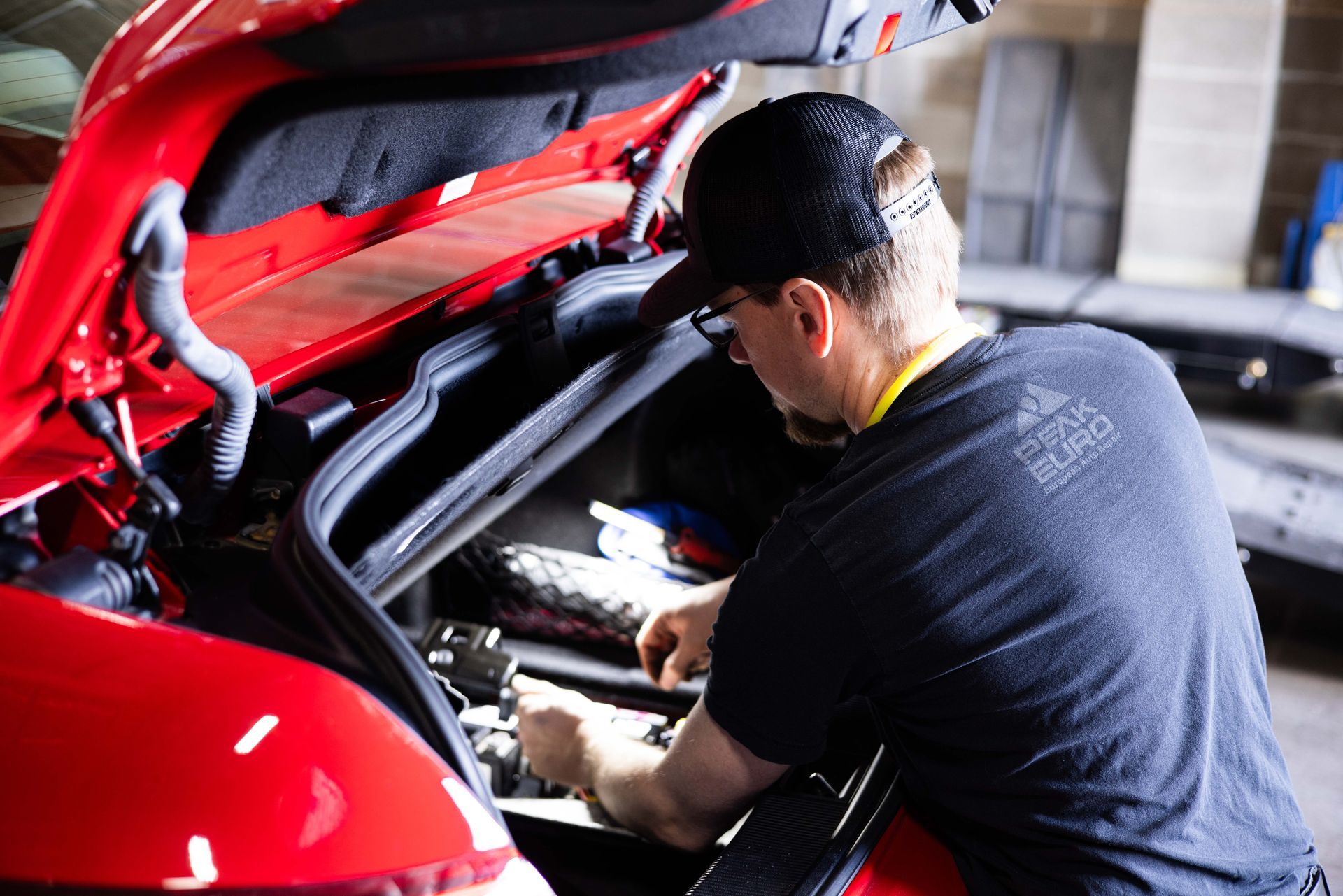 Mechanic working on a red car's engine bay. Wearing glasses, a cap, and a gray shirt. Workshop setting. | Peak Euro