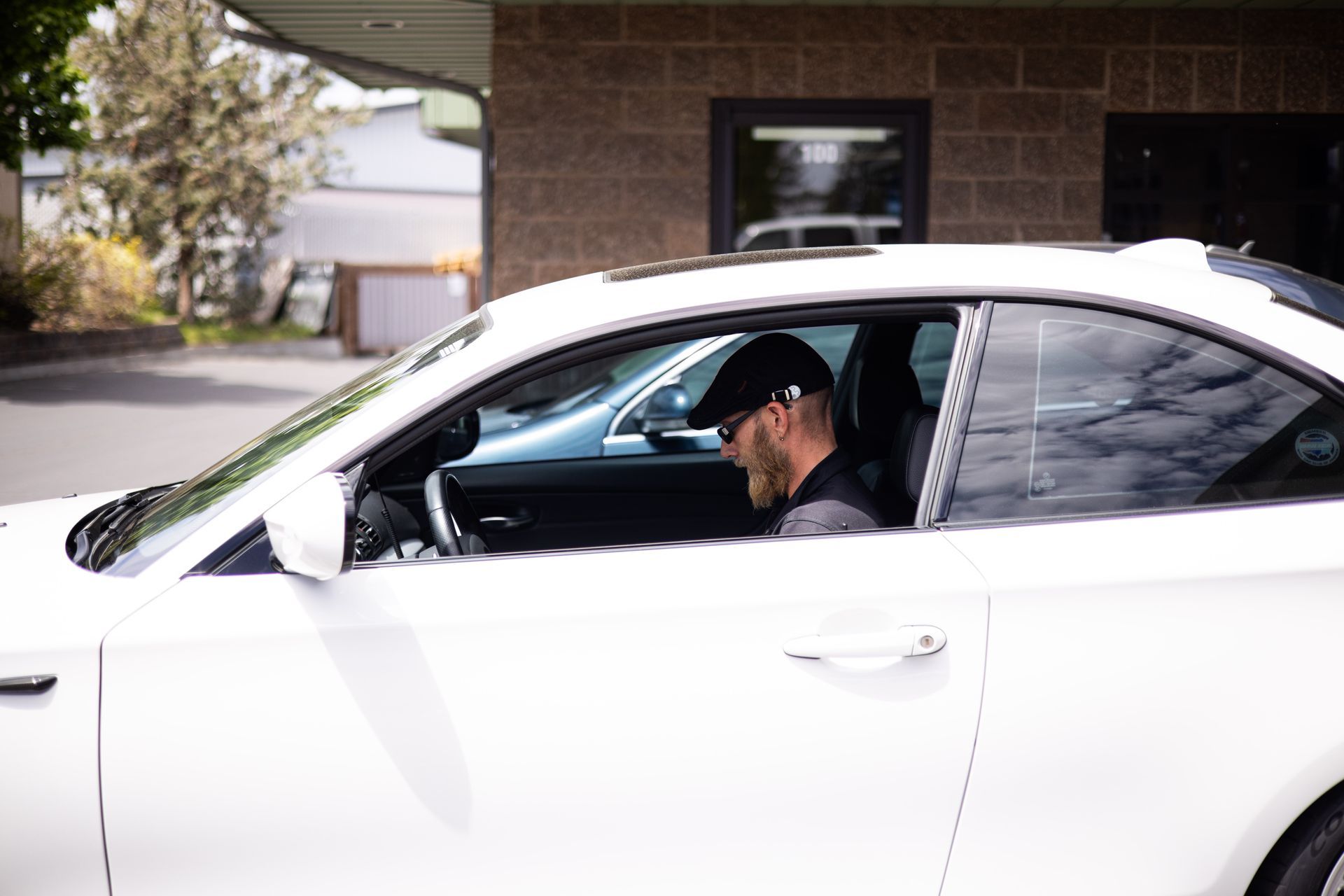 Man in cap driving white car outside a brick building.