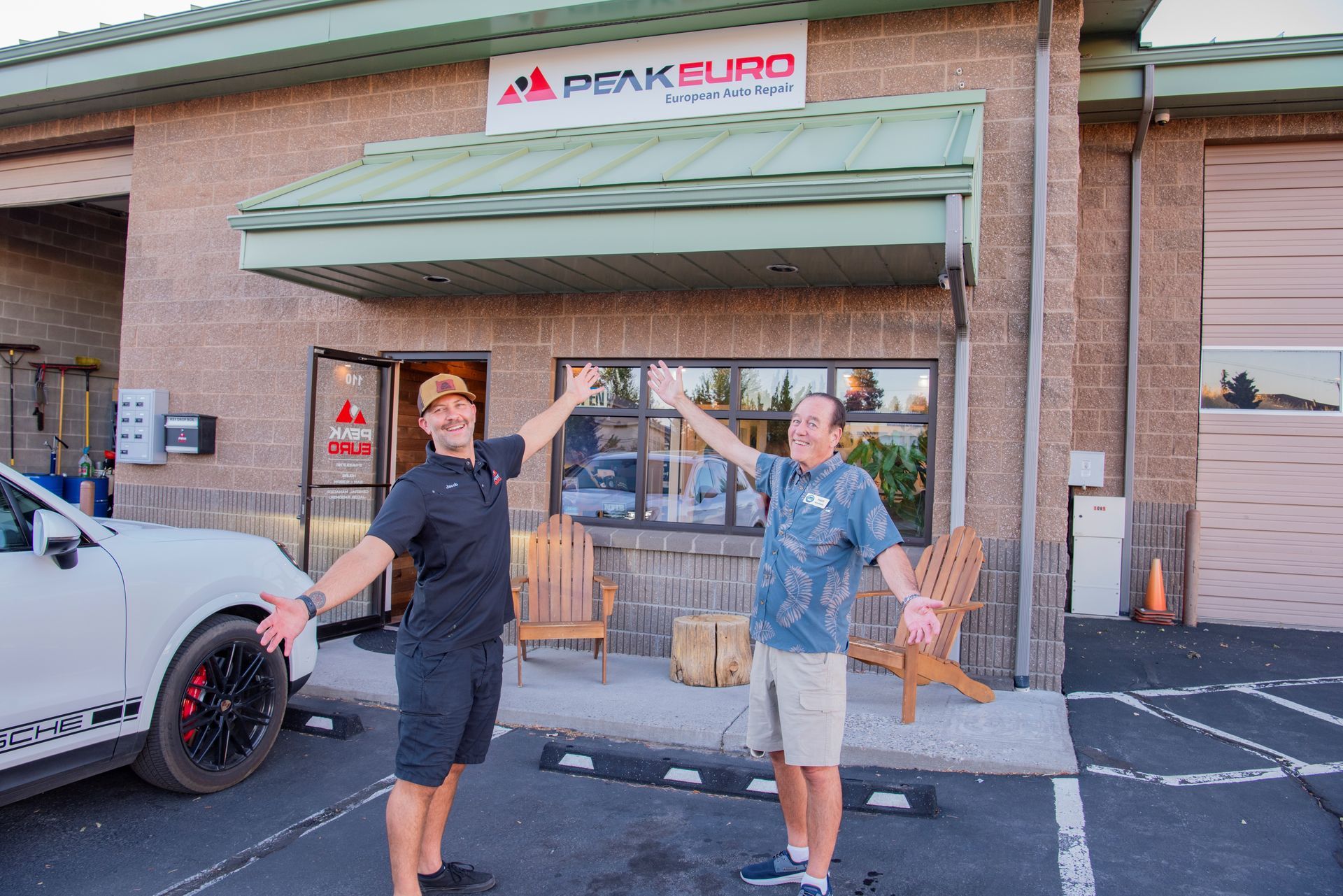 Two men with arms up in front of Peak Euro shop. White car, brick building, sunny.| Peak Euro