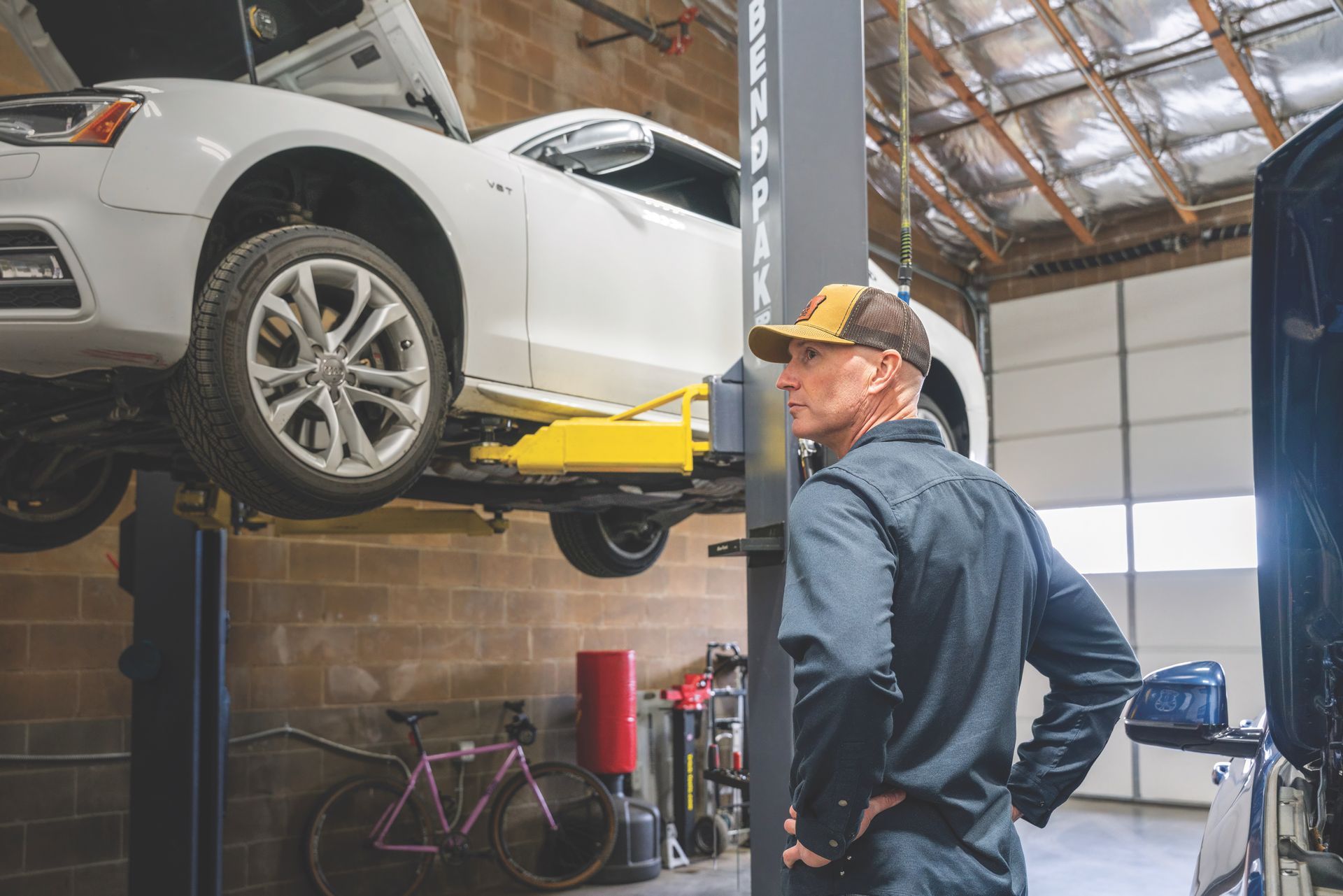 Mechanic in a garage looking at a white car on a lift. He's wearing a hat and work uniform.| Peak Euro