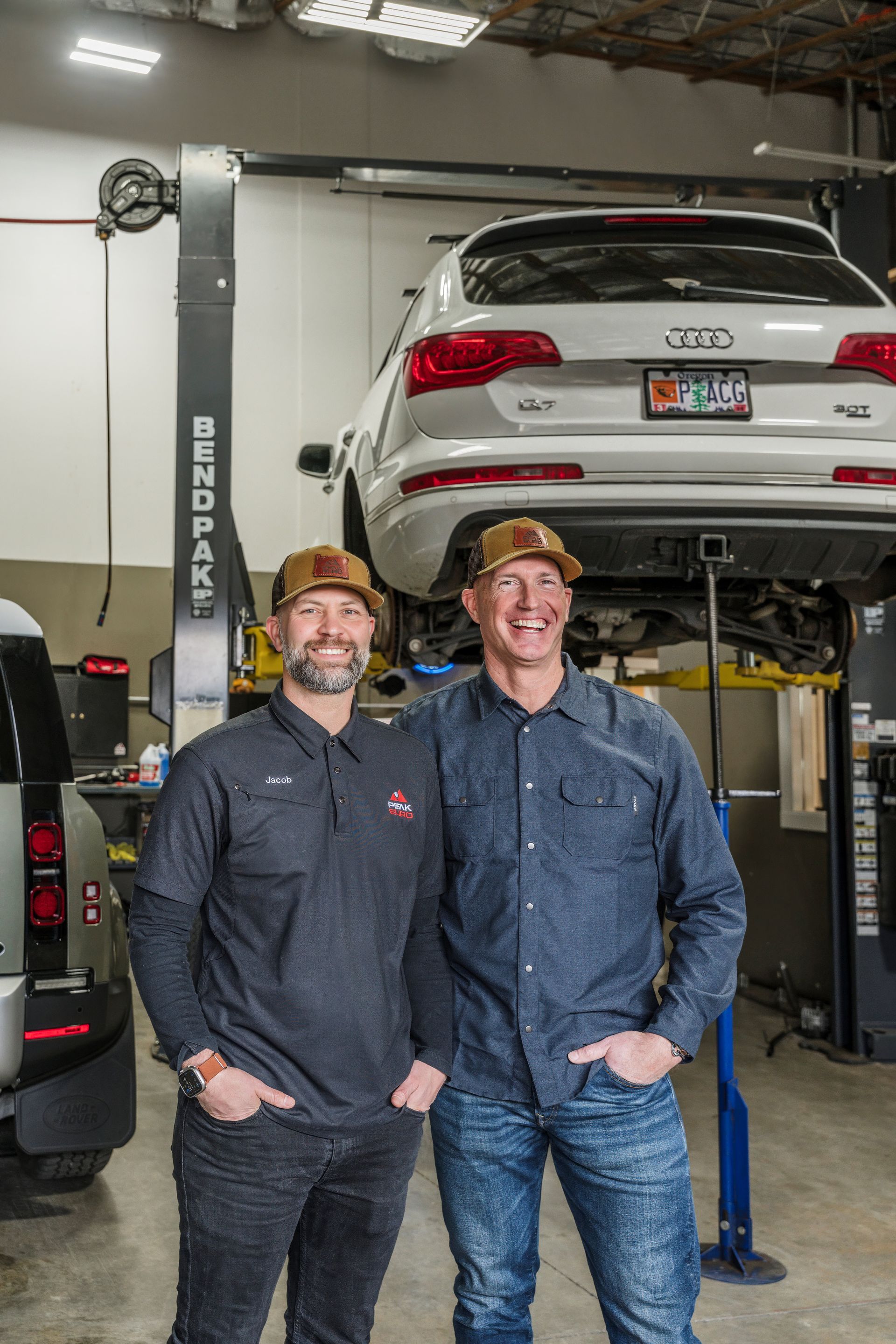 Two men pose in an auto shop. An Audi is on a lift. Both men wear hats and smile.| Peak Euro