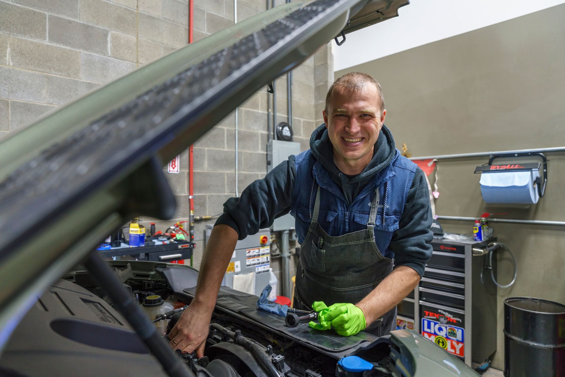 Mechanic smiling while working on a car engine in a garage; wearing gloves and apron.| Peak Euro