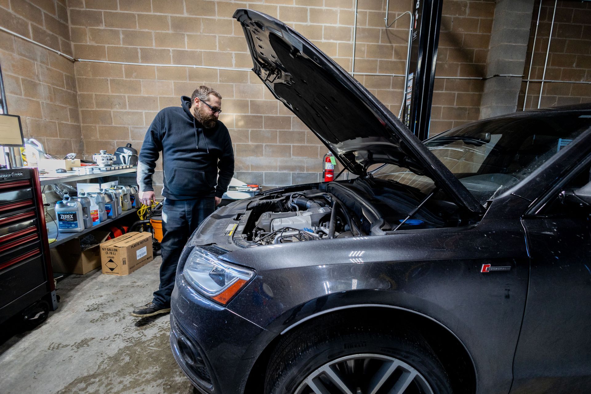 Mechanic inspecting car engine in a garage. Dark-haired man wearing glasses and a black hoodie.| Peak Euro