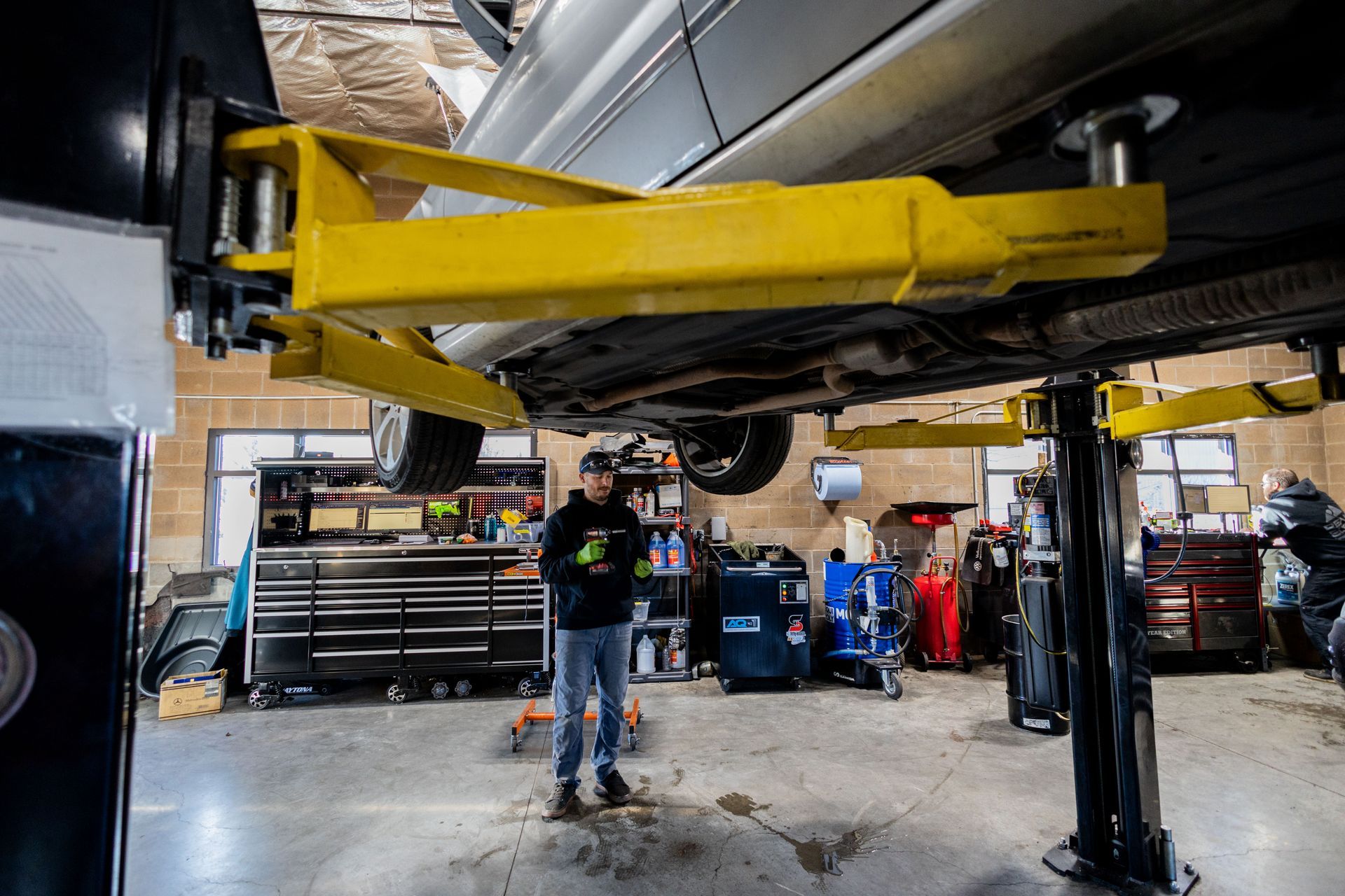 Man in a garage beneath a car on a yellow lift, surrounded by tools.