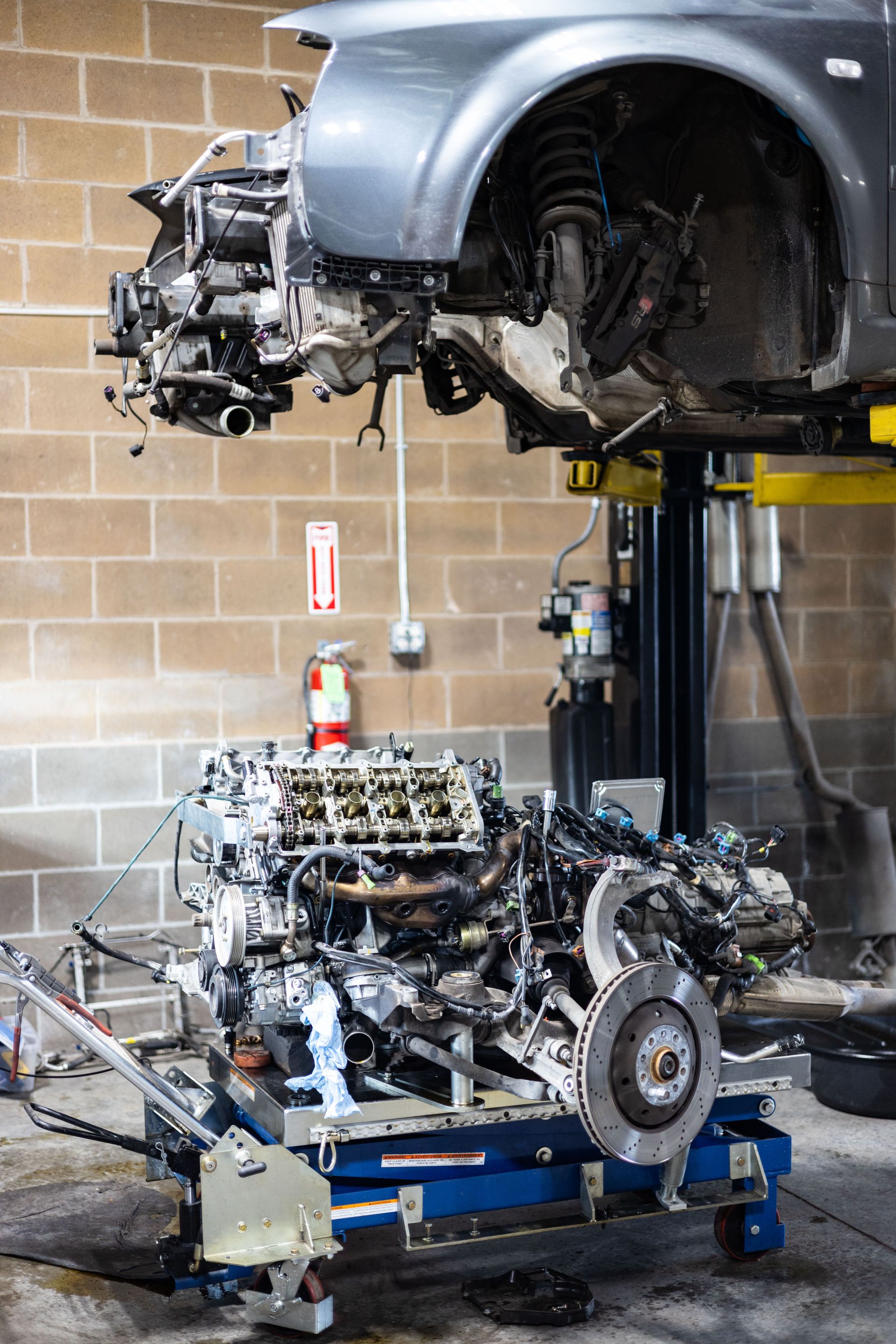 Car being worked on in a garage. Engine removed and on a cart below the car.| Peak Euro