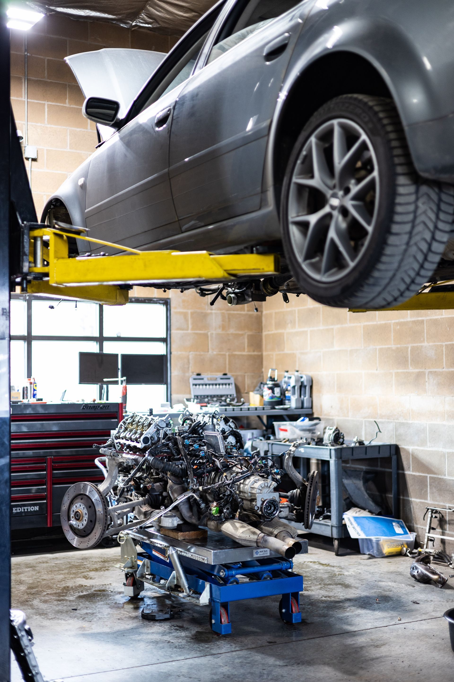 Car raised on a lift in a shop, engine on a cart below.  A mechanic's workspace with tools is in the background. | Peak Euro