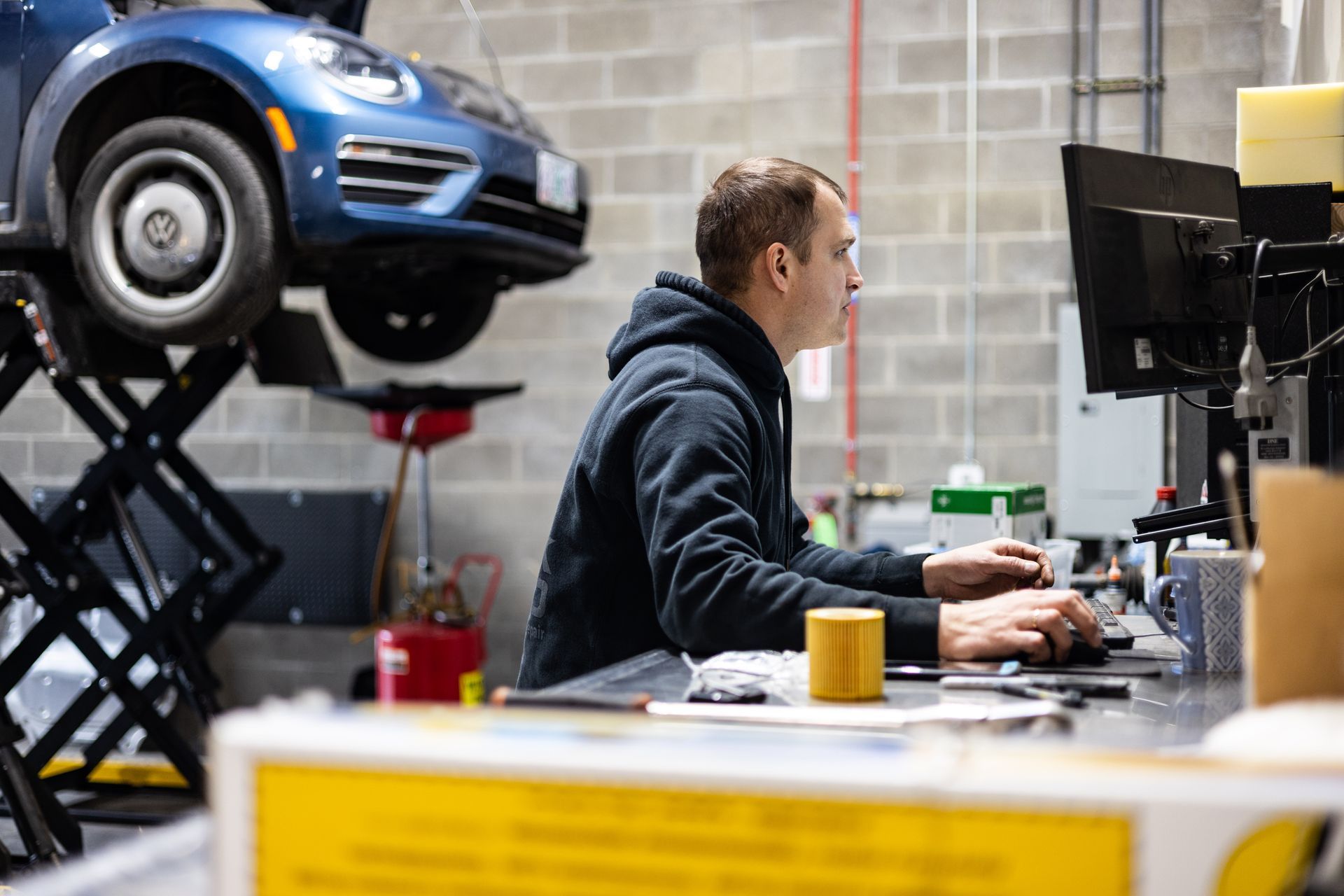 Mechanic at computer, car on lift in auto shop. Man types, blue car, brick wall. | Peak Euro