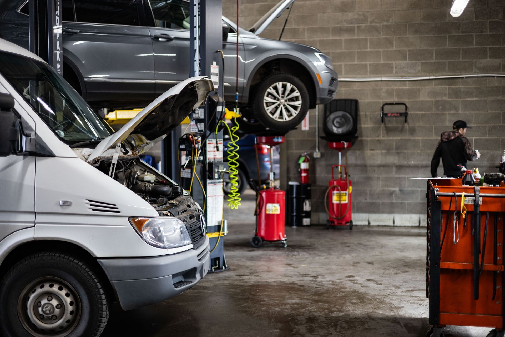 Cars being serviced in a garage. A mechanic stands near a toolbox, with a van and SUV on lifts. | Peak Euro