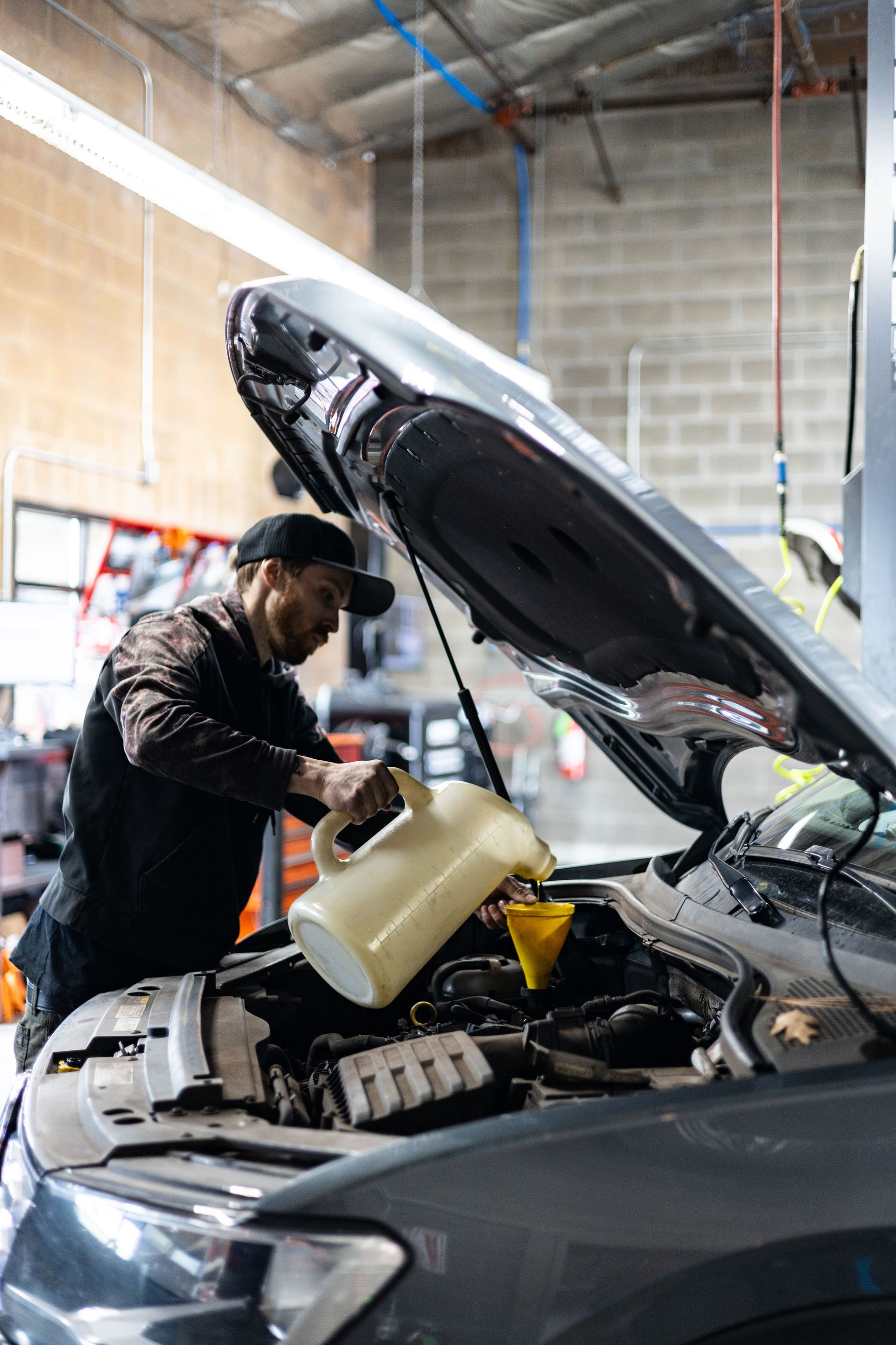 Mechanic pours oil into a car engine in a garage. He wears a black hat and jacket. | Peak Euro