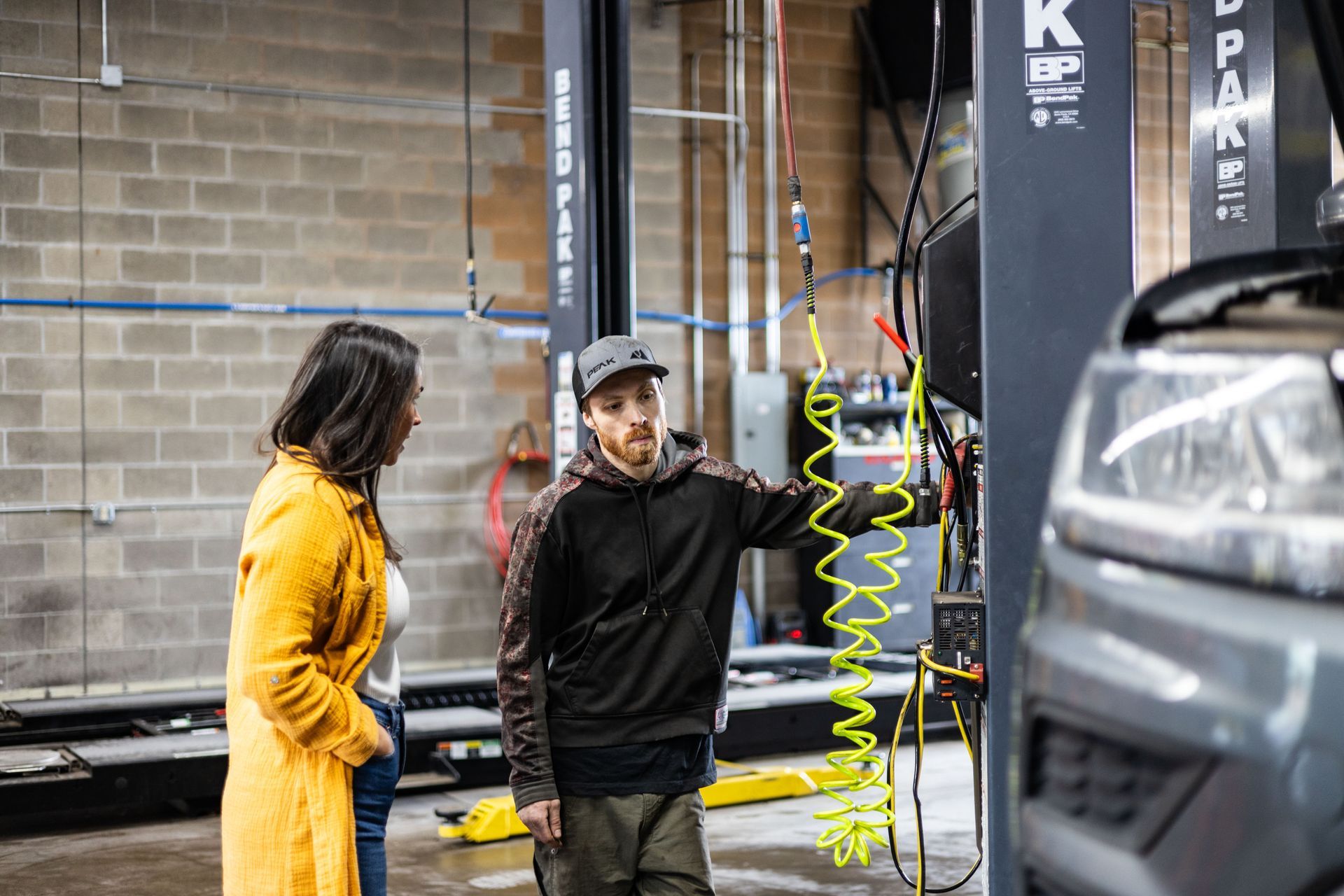 Mechanic explains car diagnostics to a woman in a garage. The mechanic wears a hoodie and hat. The woman wears a yellow coat. | Peak Euro