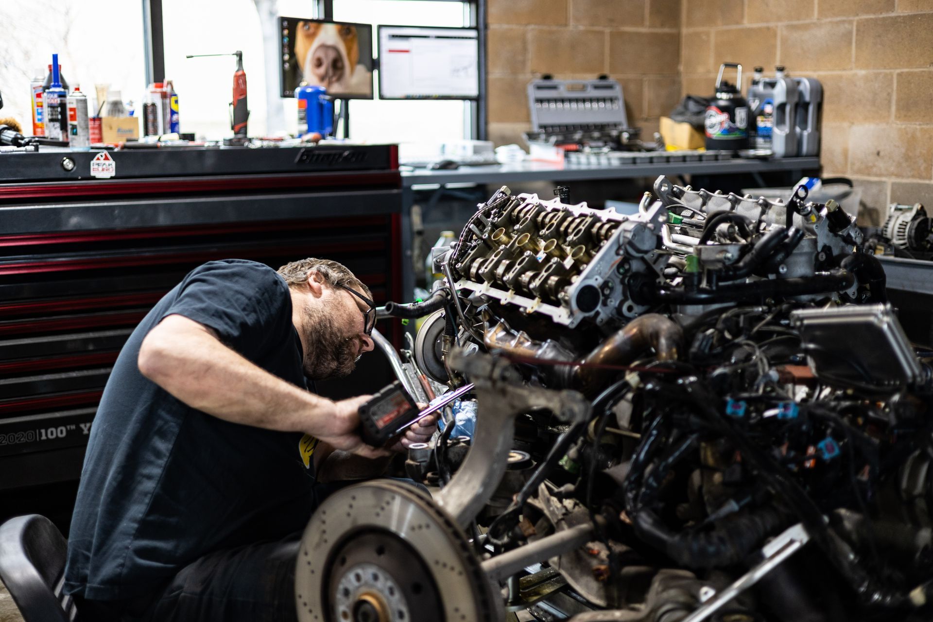Mechanic works on car engine in a garage. He is wearing sunglasses, and the engine is partially disassembled. | Peak Auto