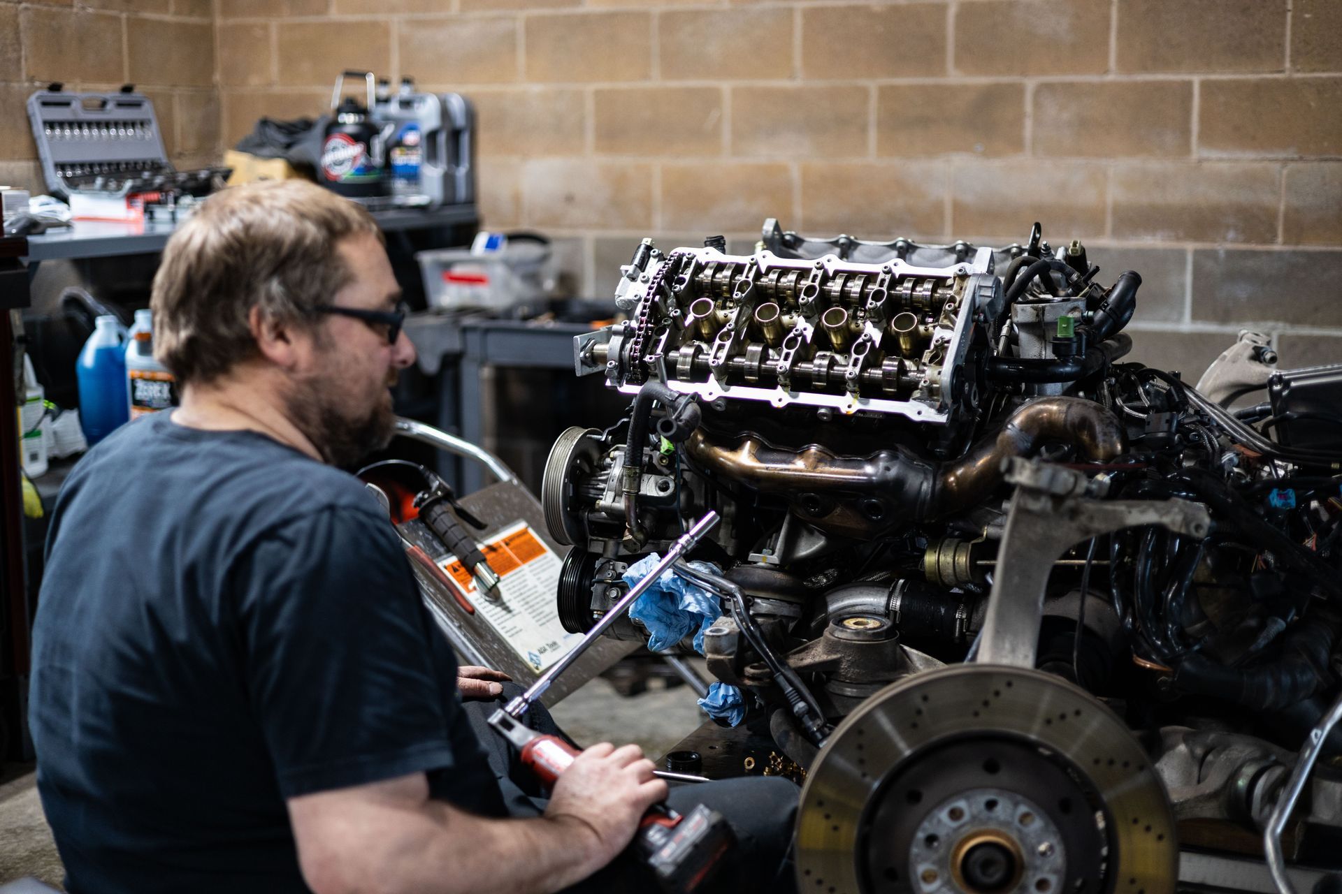 Mechanic works on car engine in a garage. He is wearing sunglasses, and the engine is partially disassembled. | Peak Auto