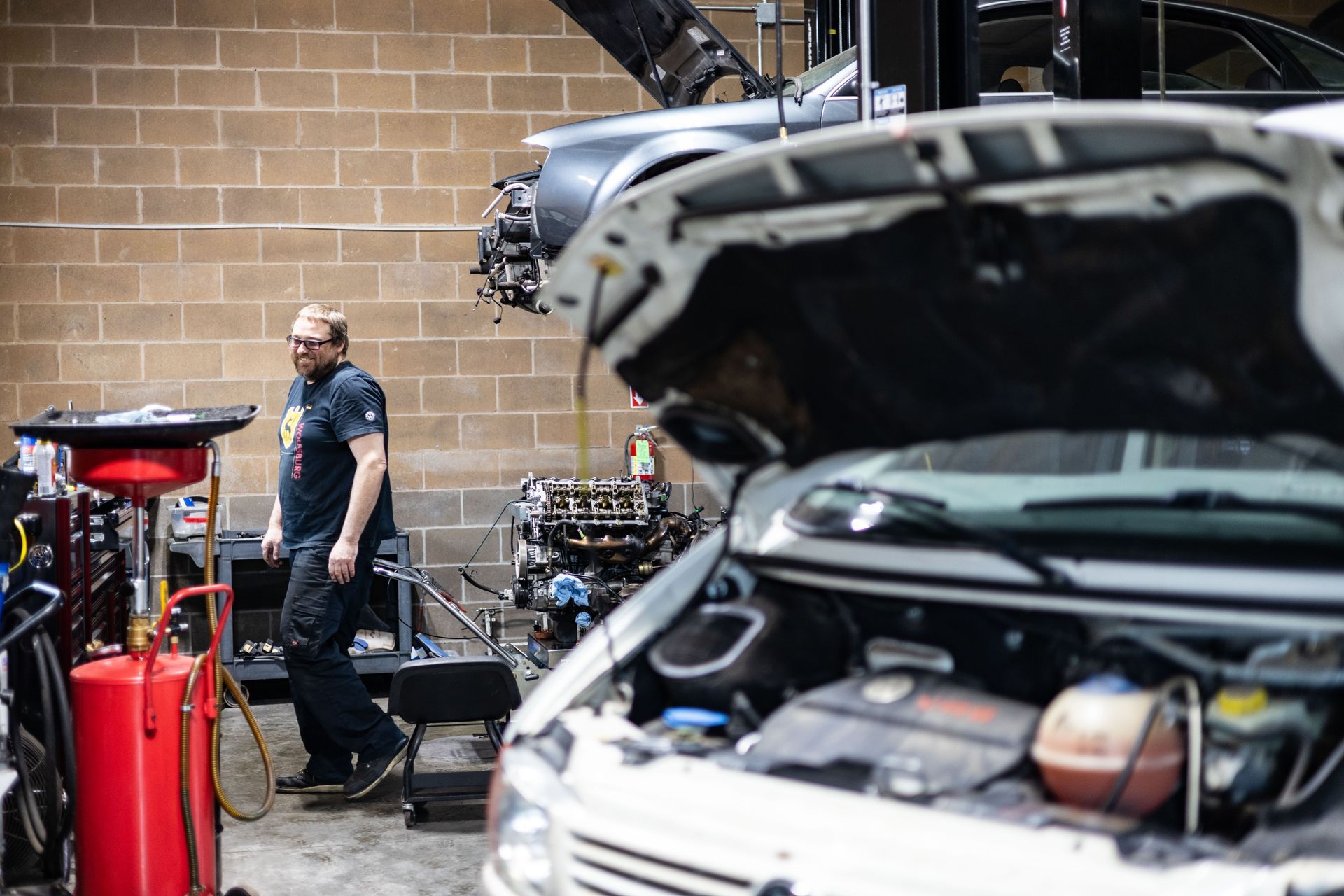 Mechanic in a garage, standing near a car with its hood open, other car parts visible, brick wall background.