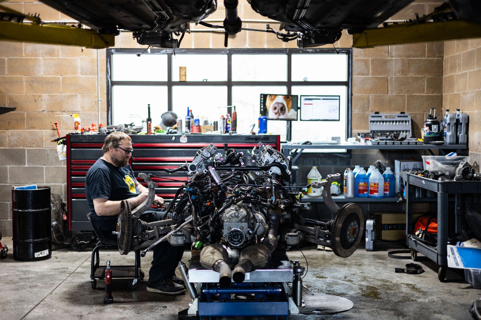 Mechanic working on a large car engine inside a garage, with tools and parts visible.