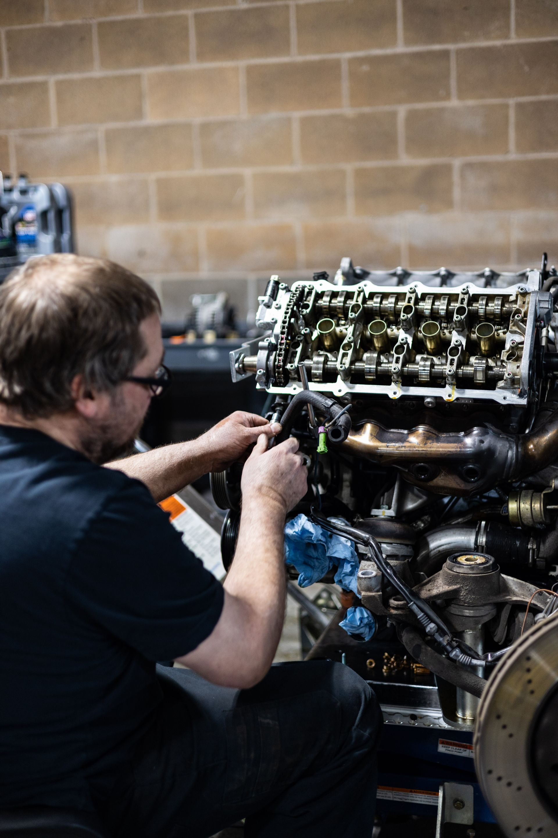 Mechanic works on a car engine in a brick-walled garage, focusing on internal components.