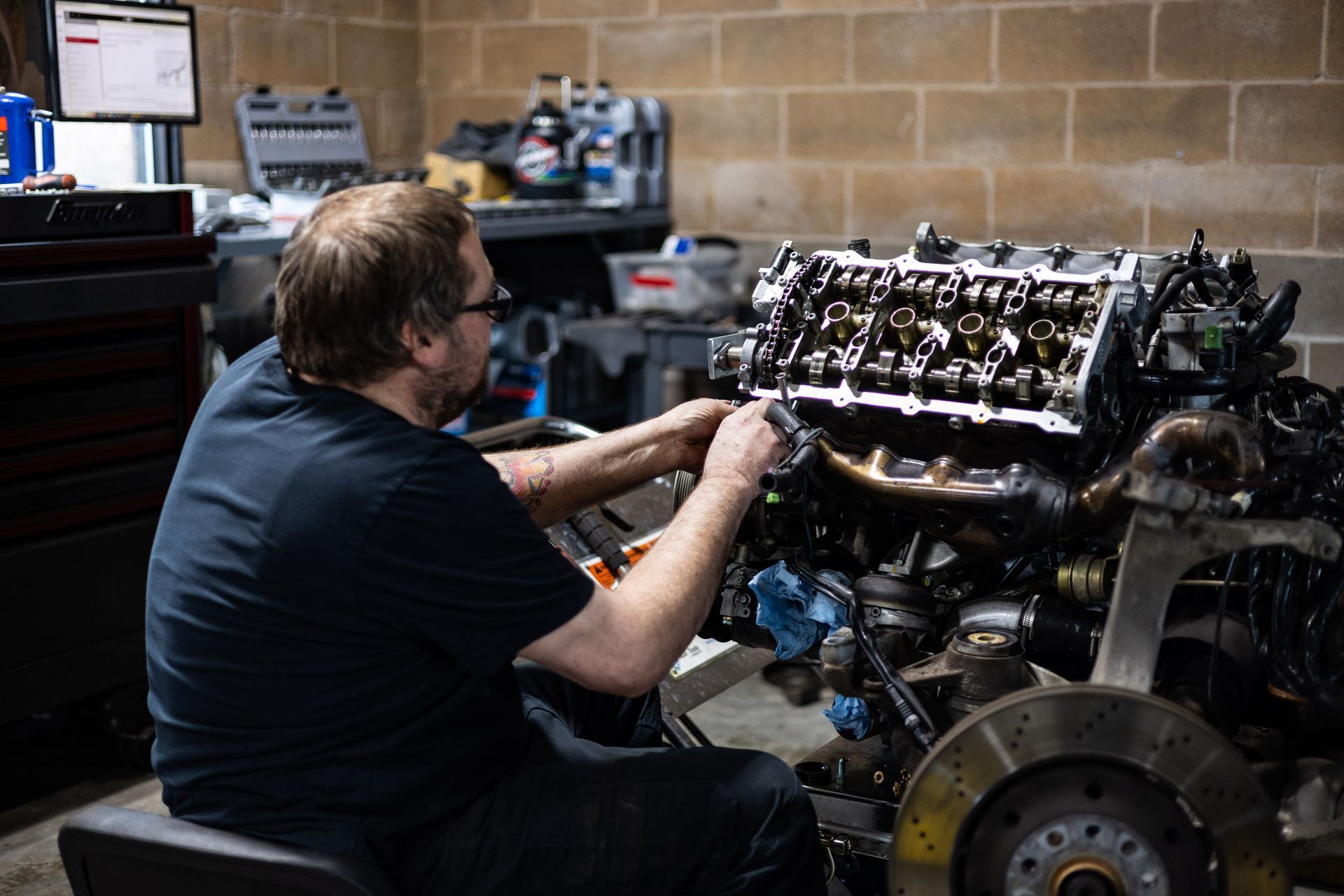 Mechanic working on an engine in a garage, wearing glasses and a black shirt, brick wall background.