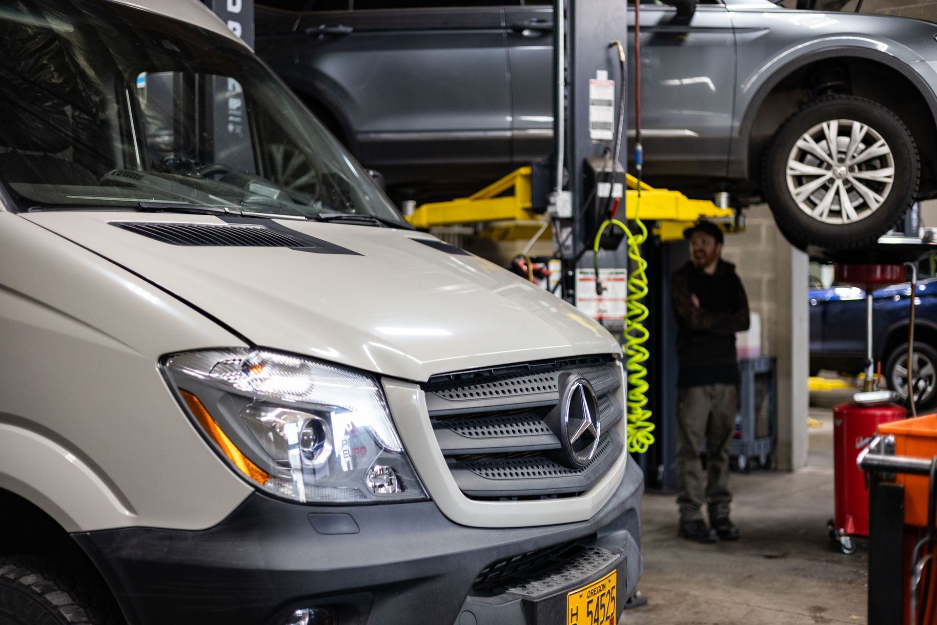 Mercedes van in auto shop, man standing nearby, car on lift above. | Peak Euro