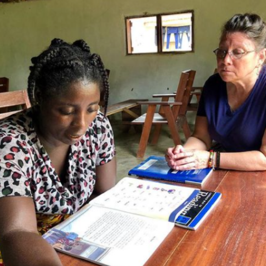 Two women seated at a table, studying an open book. One wears a patterned shirt, the other a blue top.