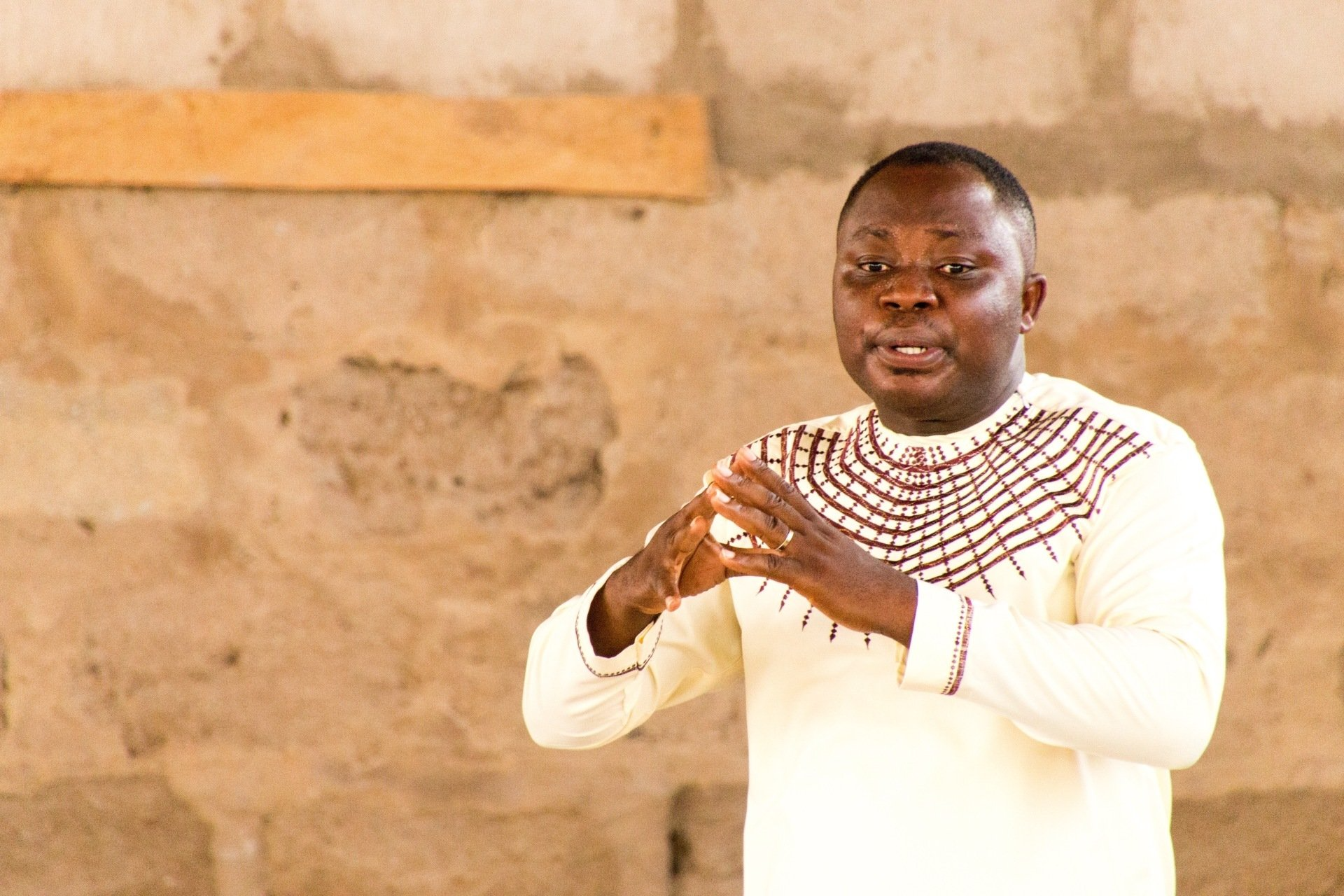 Man in light-colored shirt gestures with hands, speaking, against a textured, light-brown wall.