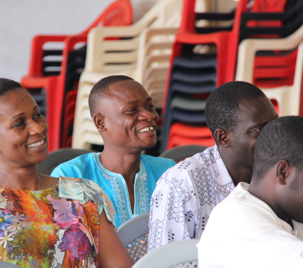 People smiling in a room; colorful plastic chairs stacked in the background.