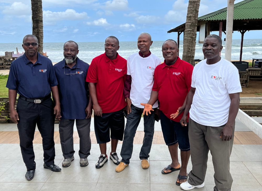 Group of six men posing outdoors near a beach, wearing various colored shirts and pants.