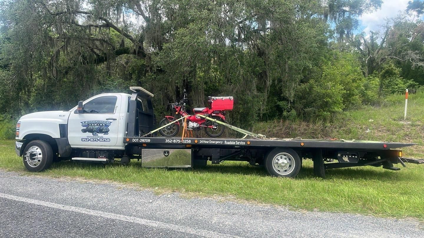 Tow truck with a red motorcycle secured on the flatbed, parked on a grassy roadside.