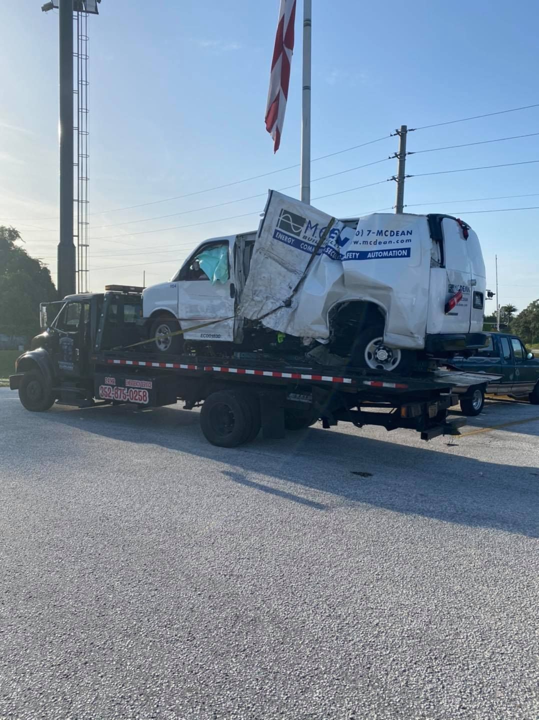A wrecked white van on a tow truck bed, damaged front and side, parked by a building, flag overhead.