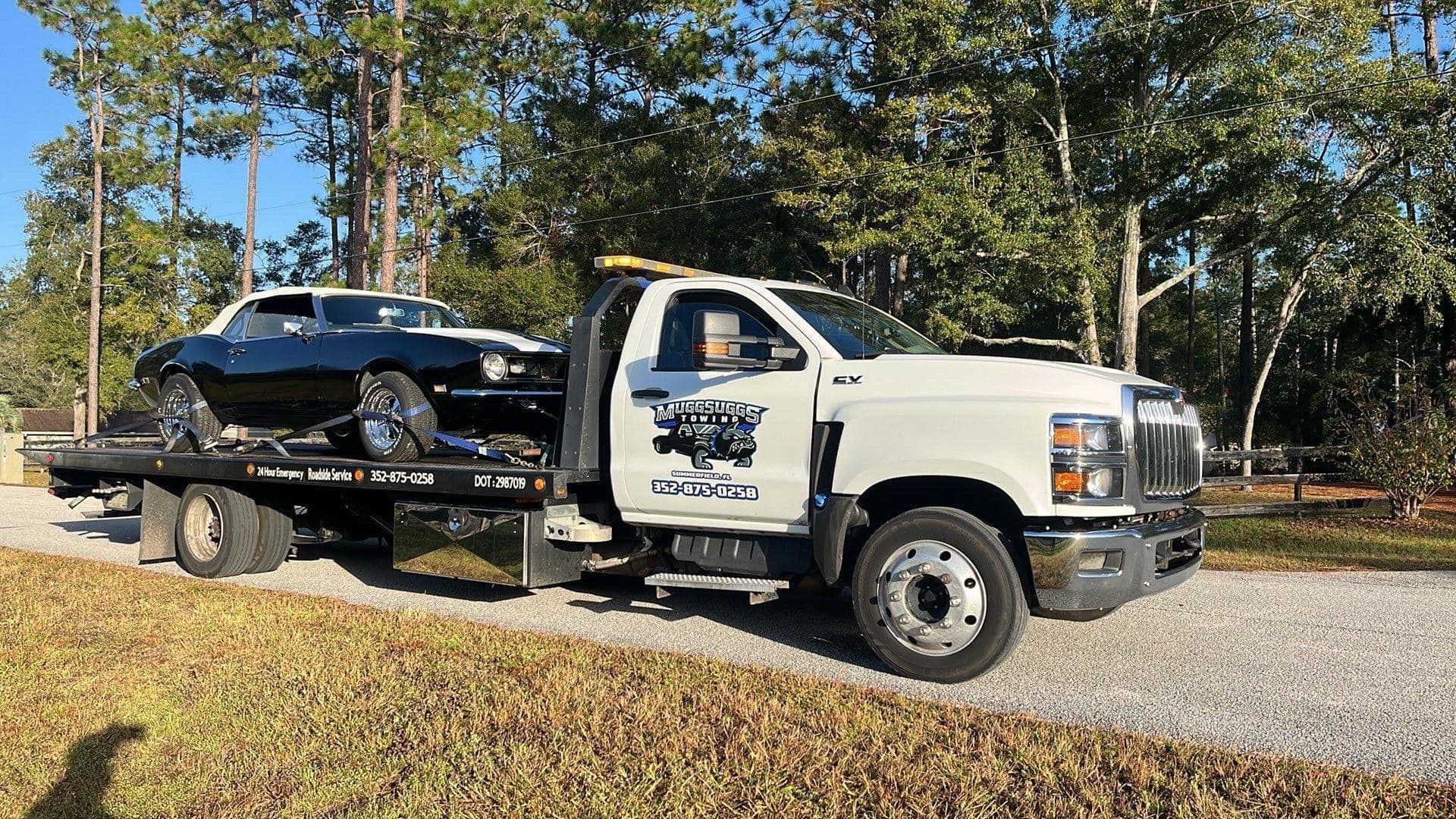 A black classic car being towed by a white tow truck on a gravel road, trees in the background.