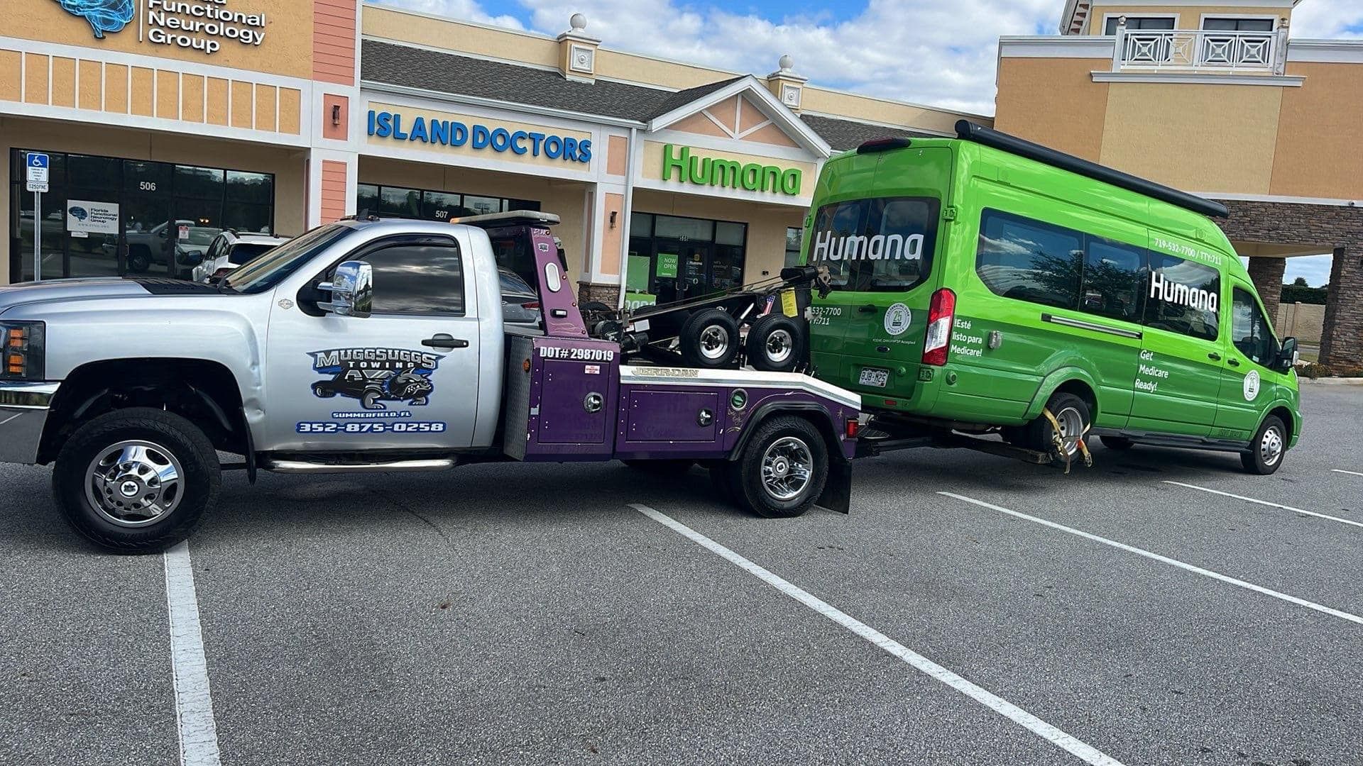Tow truck towing a green van in front of a shopping center.