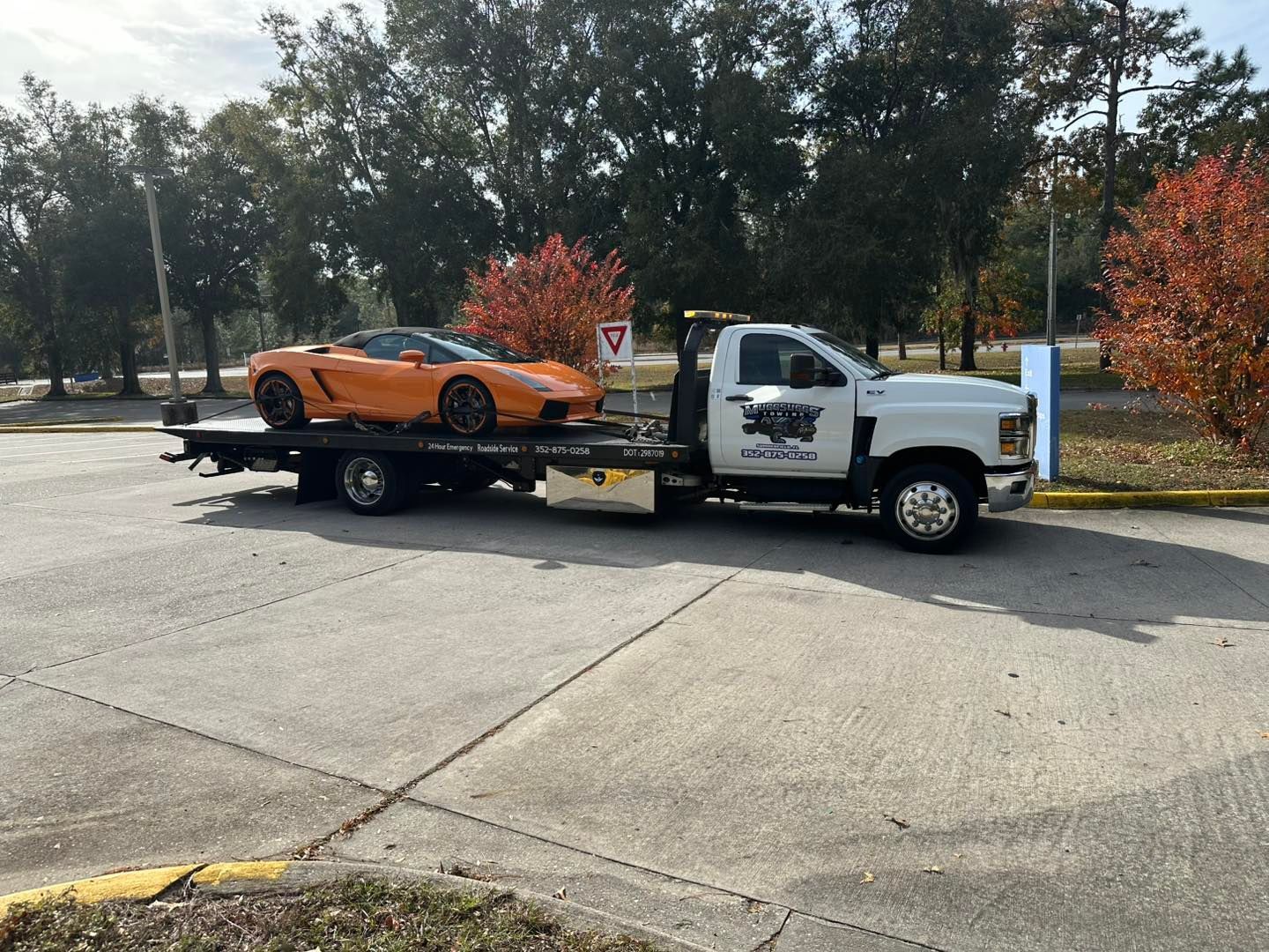 Orange sports car on a flatbed tow truck, parked on a paved surface near trees.