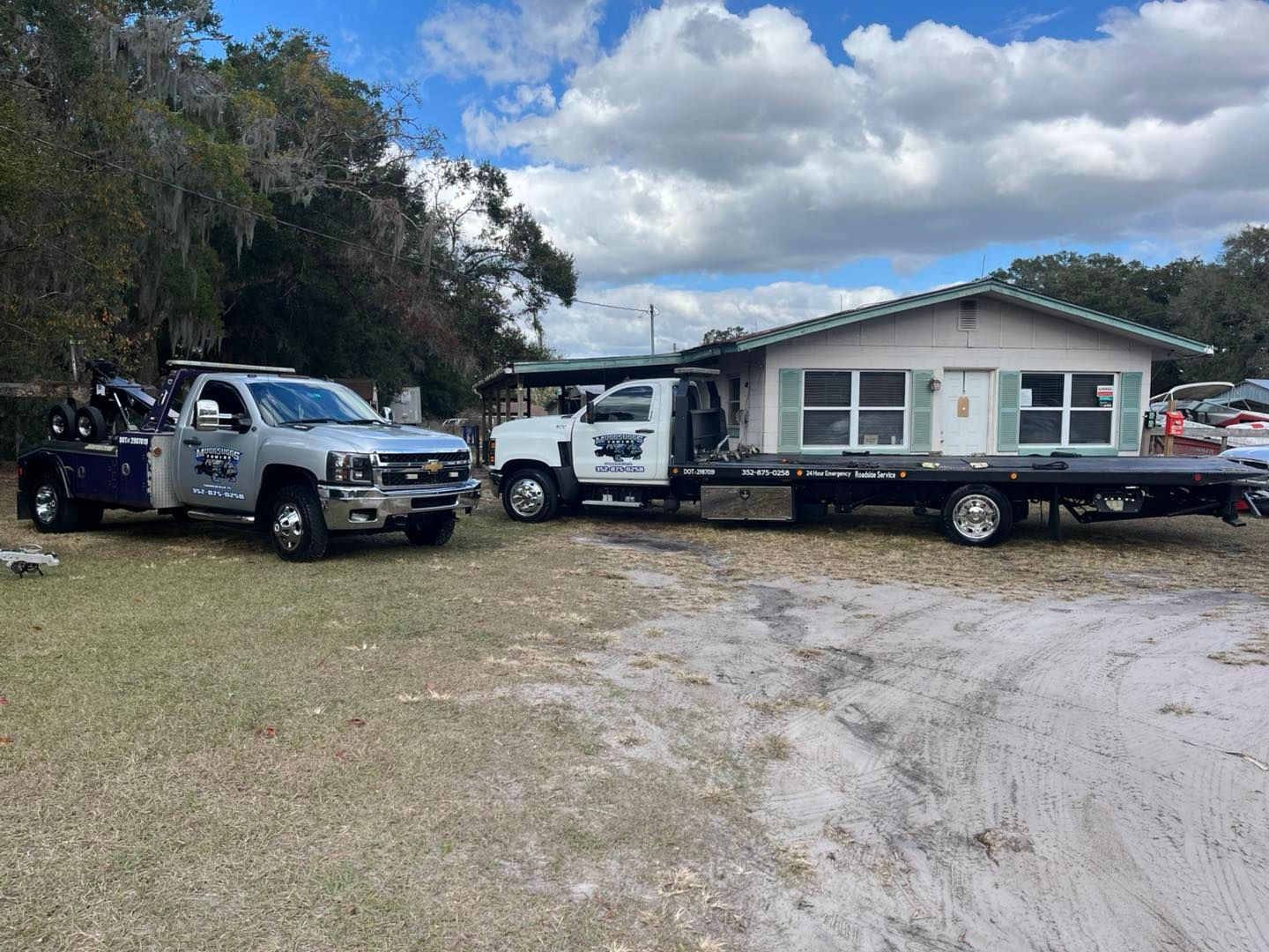 Two tow trucks in front of a small house. One is blue/silver, the other white, both with logos.