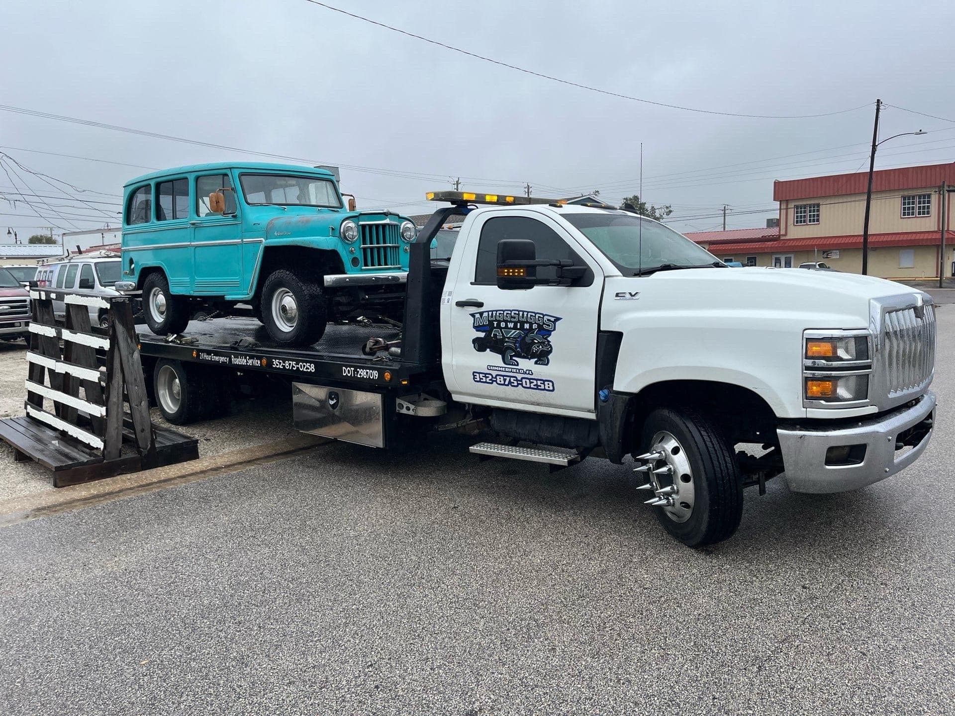 A turquoise Jeep wagon is on a white tow truck, in front of a building on a rainy day.