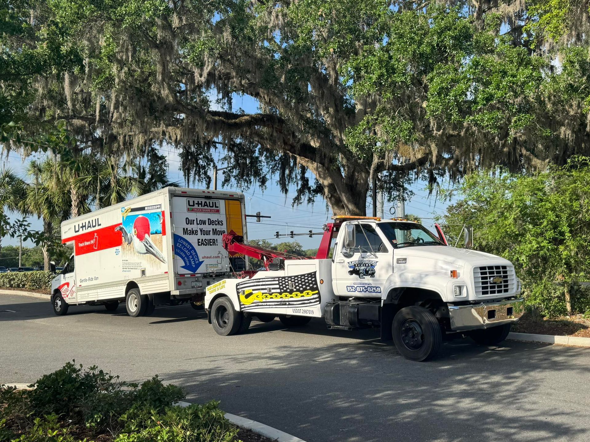 A white tow truck towing a U-Haul truck under a large tree; sunny outdoor setting.