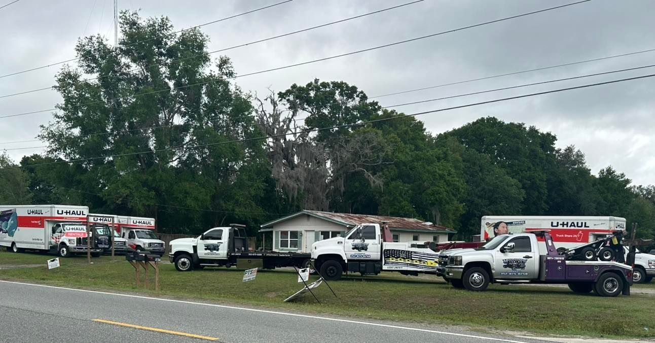 U-Haul trucks and tow trucks parked in front of a house on a grassy lot. Overcast sky.
